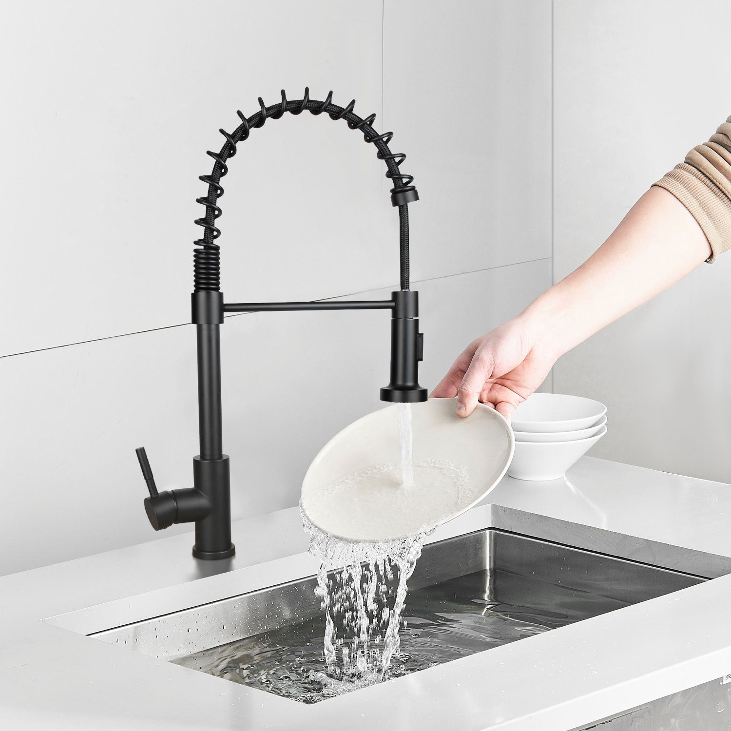 Czarny kran kuchenny spryskuje wodą talerz w zlewie ze stali nierdzewnej. Osoba trzyma talerz. W pobliżu są białe miski.