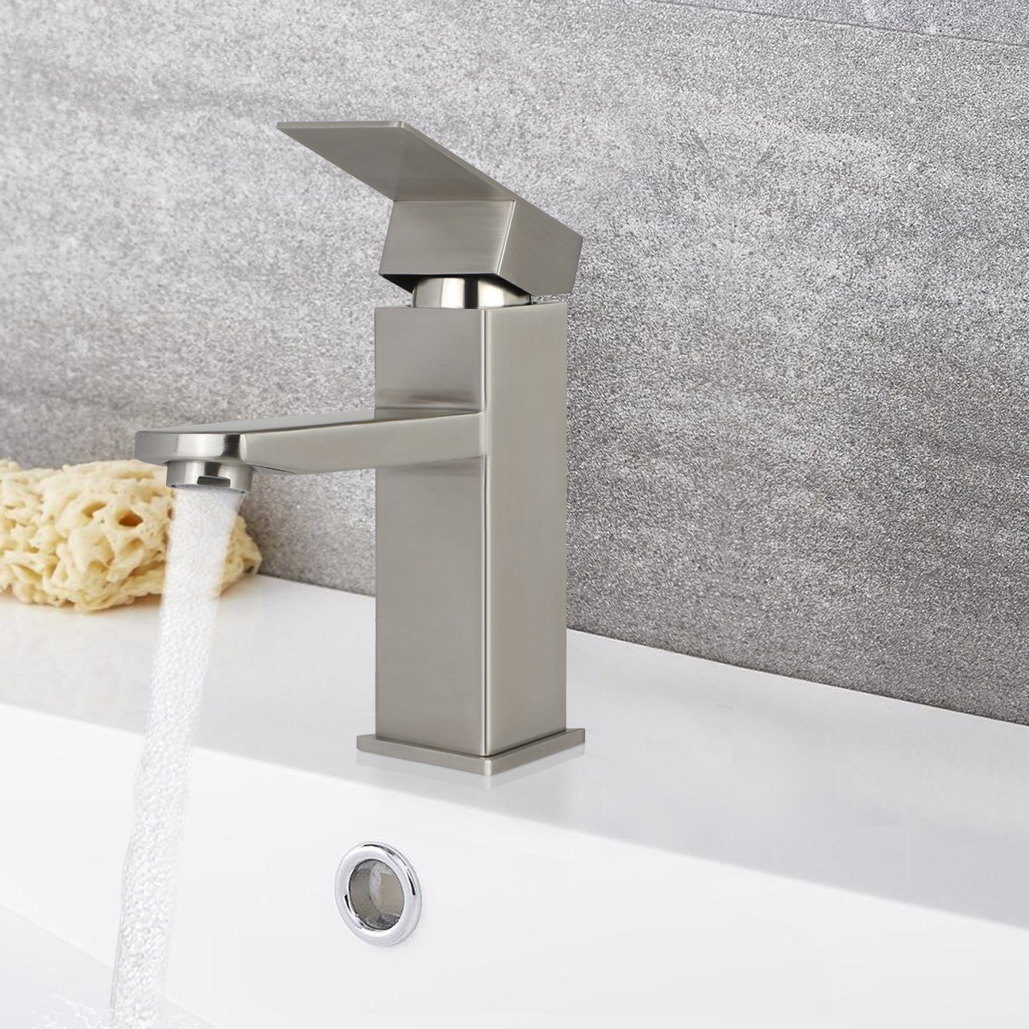 Nowoczesna srebrna bateria z wodą wpadającą do białej umywalki. Obok leży beżowa gąbka na tle szarej ściany.