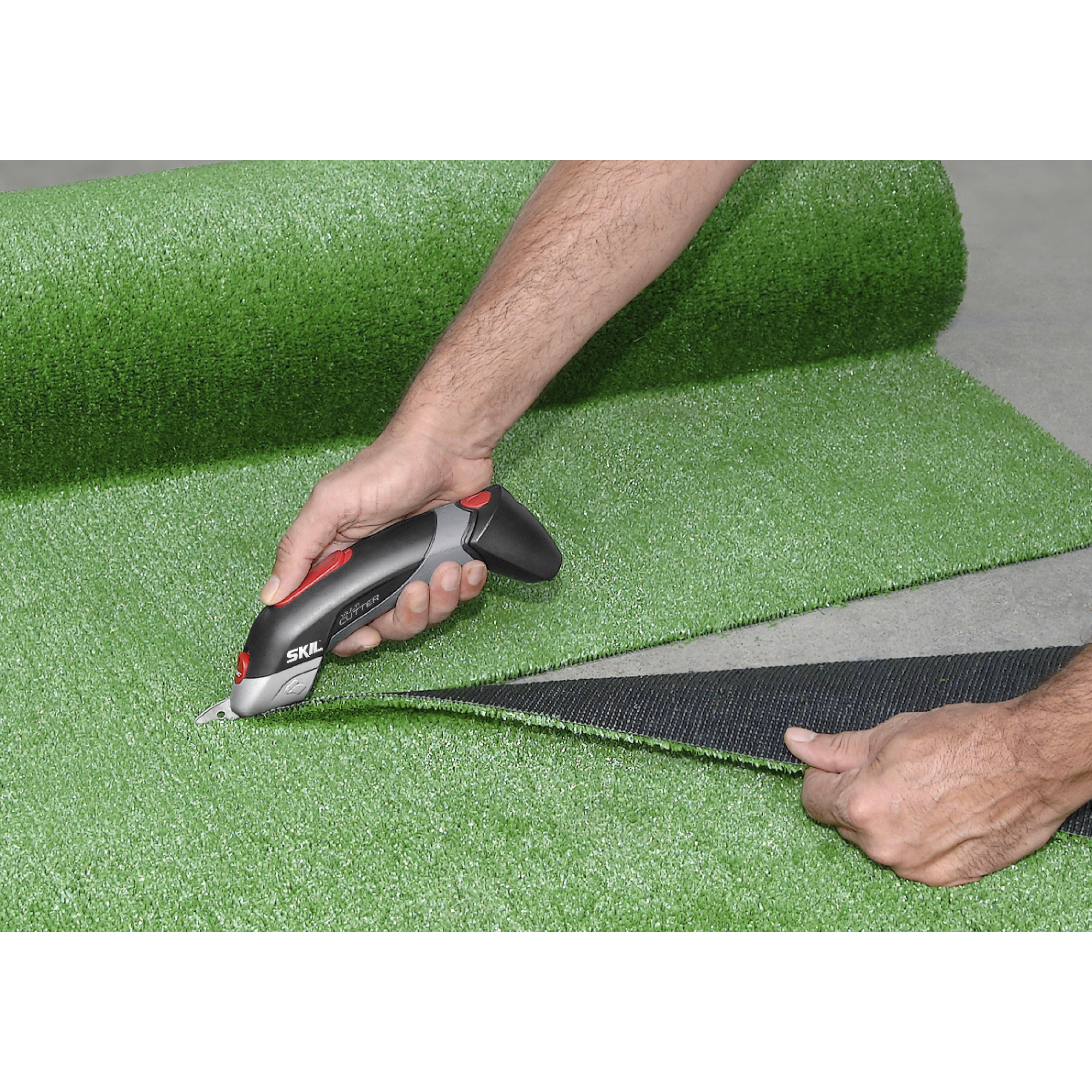 Een persoon gebruikt een SKIL-snijder om kunstgras te trimmen. Het groene gras ligt op een betonnen oppervlak. De snijder is zwart, grijs en rood.