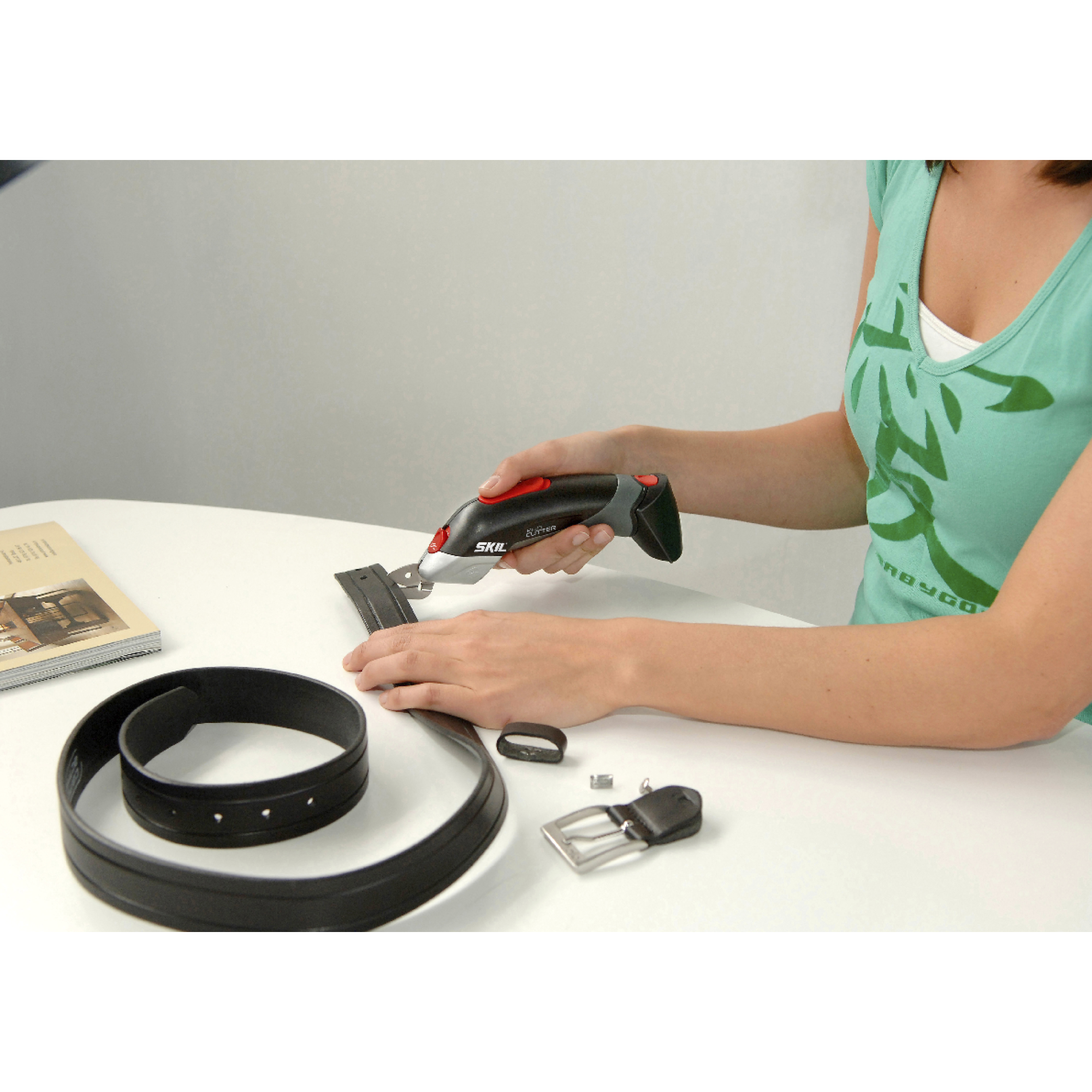 Een vrouw gebruikt een SKIL-snijder om een zwarte leren riem op een witte tafel te trimmen. Verschillende riemdelen liggen verspreid.