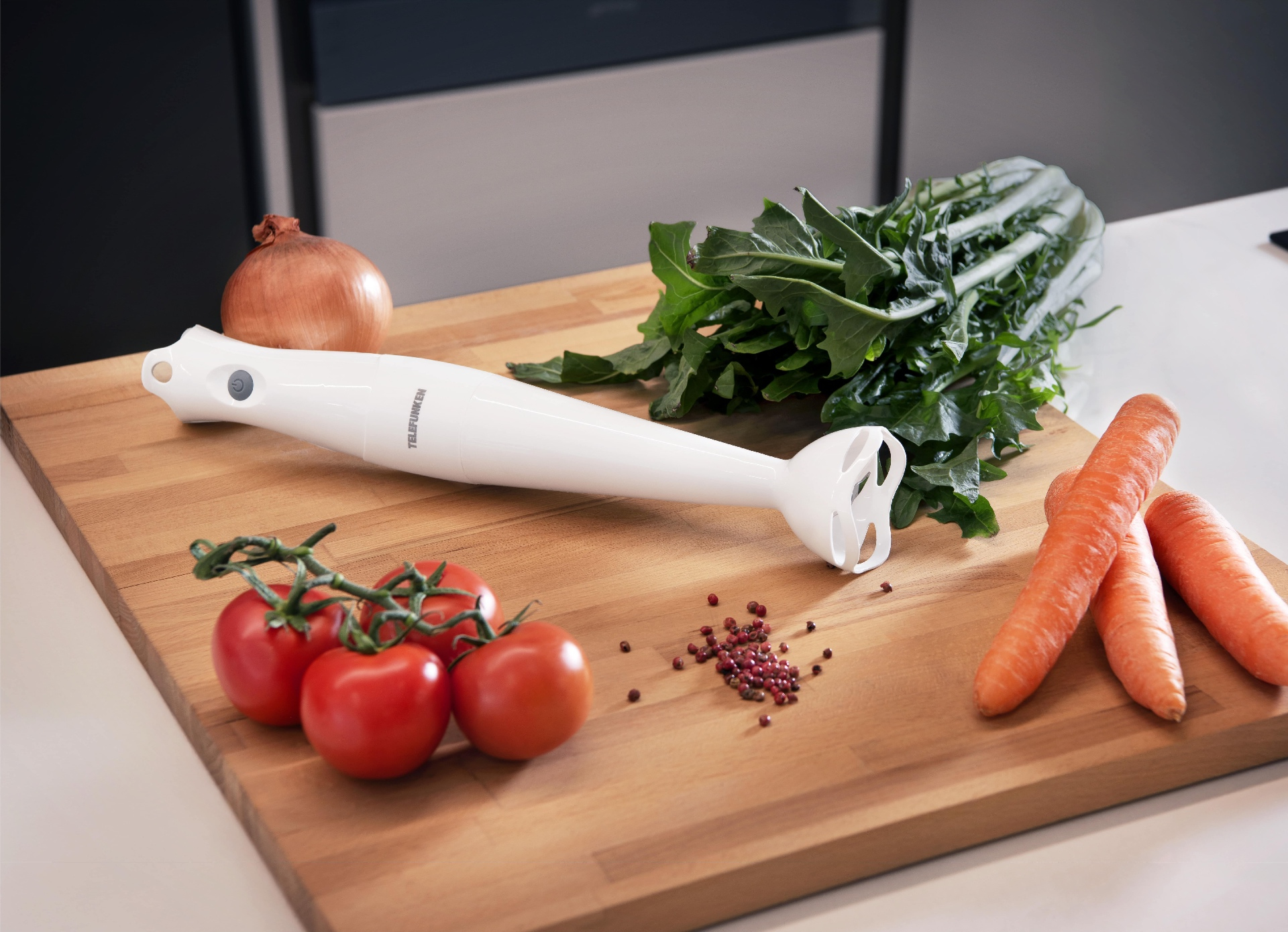 Een snijplank met een witte staafmixer, tomaten, uien, wortelen en groenten. Rode peperkorrels zijn verspreid.