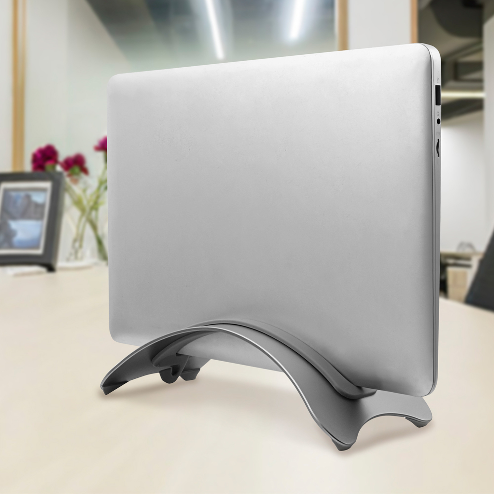 Srebrny laptop stoi na szarym stojaku na jasnym drewnianym stole. W tle rozmyte otoczenie biurowe.