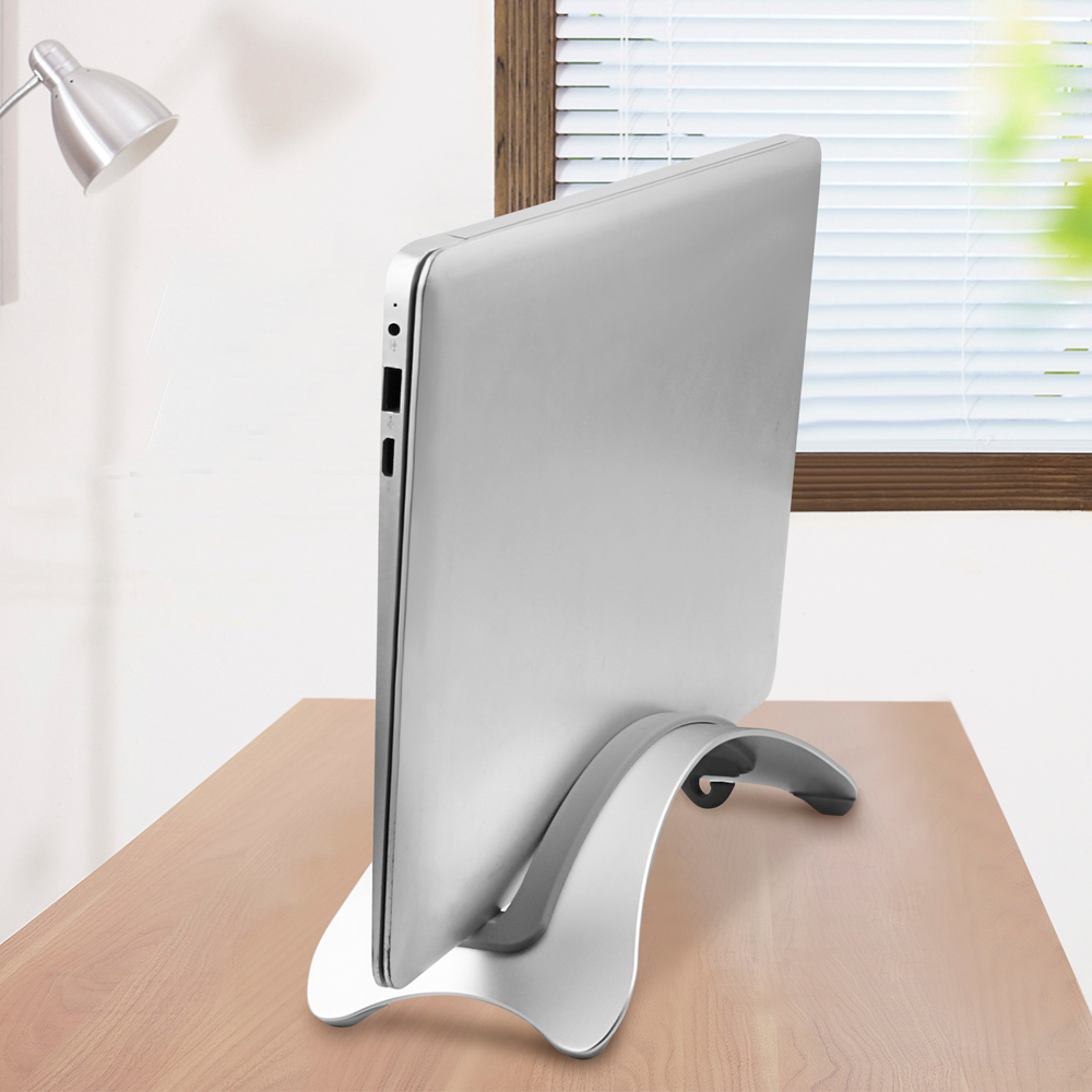 Srebrny laptop na zakrzywionym, srebrnym stojaku na drewnianym biurku. Lampa biurkowa po lewej.