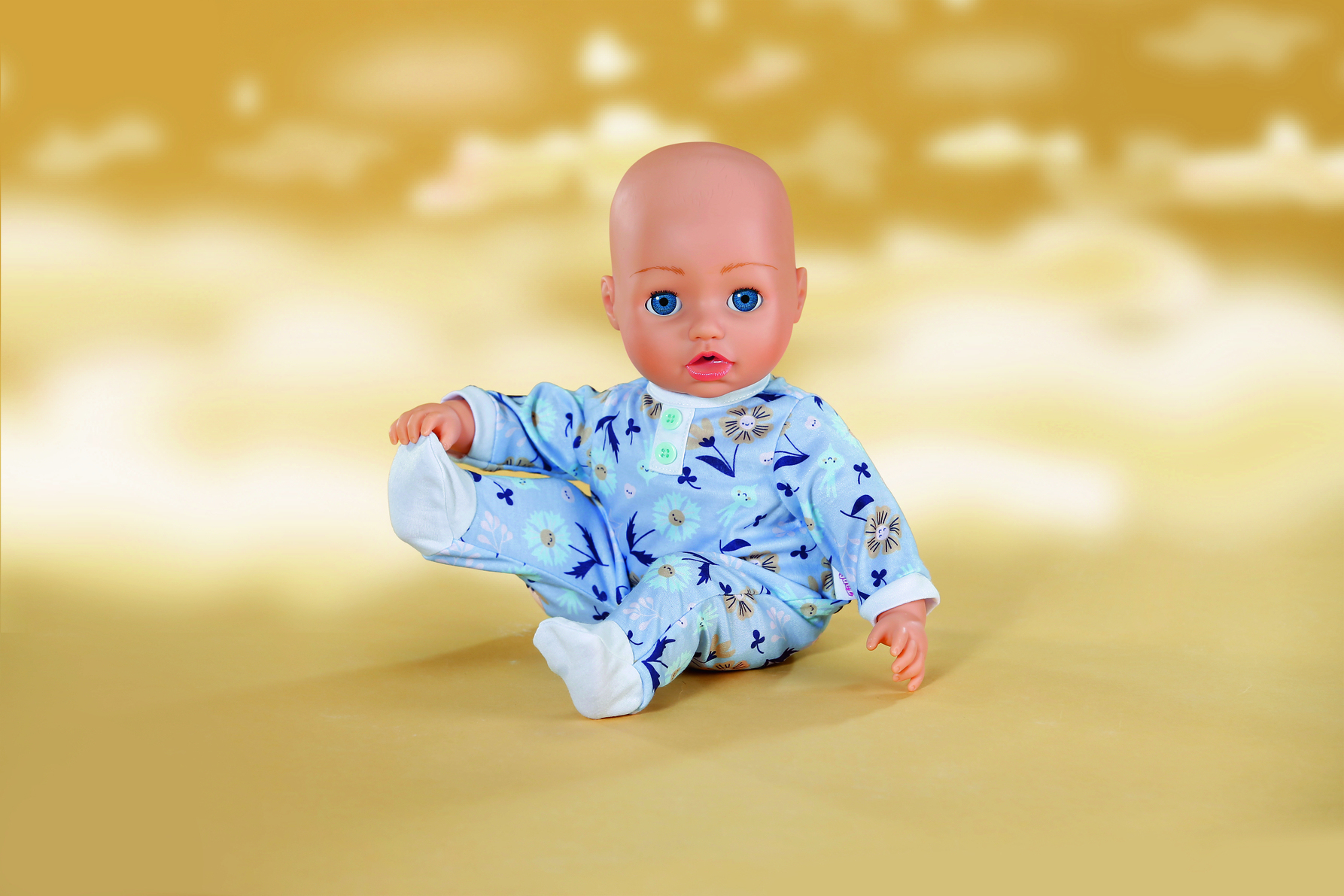 Lalka siedzi z uniesioną nogą. Ma na sobie niebieski kombinezon w kwiaty i niebieskie oczy. Tło to żółty gradient.