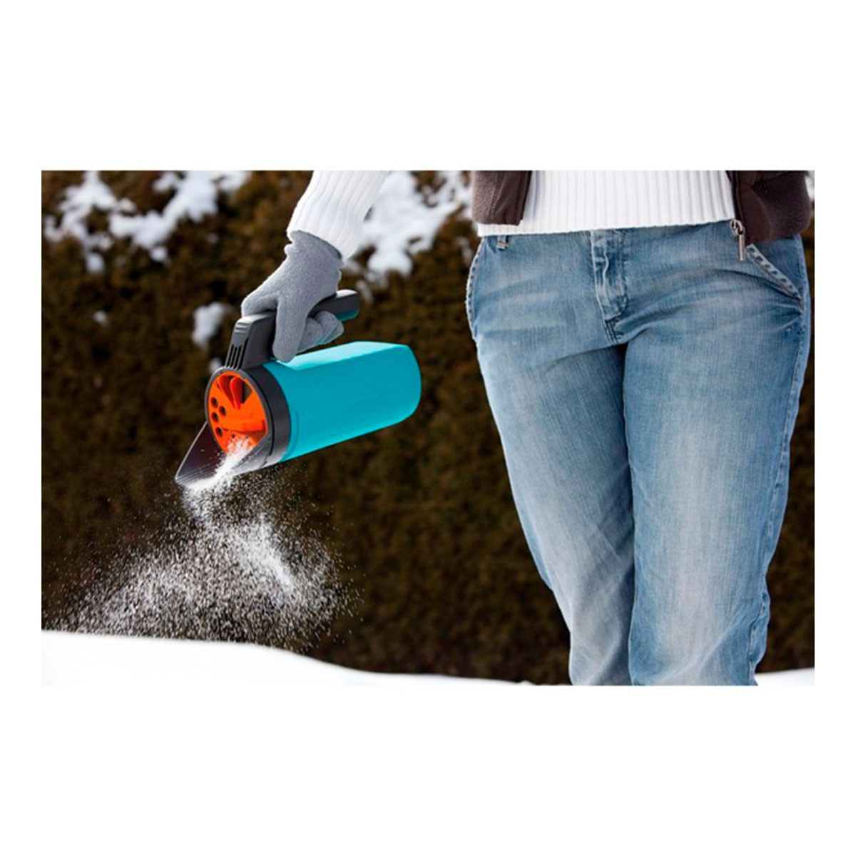 Osoba używa niebiesko-pomarańczowego rozsiewacza śniegu na zaśnieżonej powierzchni. Ma na sobie dżinsy i biały sweter. Tło to ciemnozielony krzew.