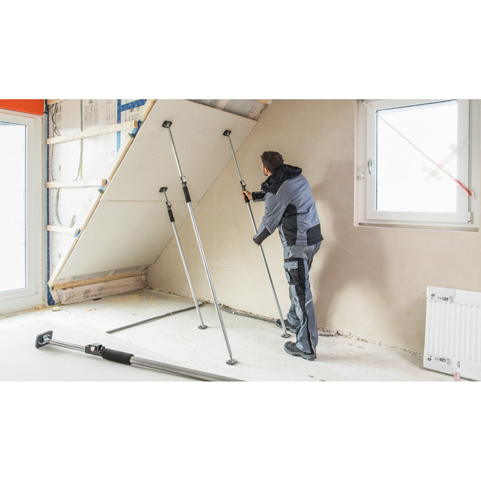 Ein Arbeiter installiert Trockenbau in einem Raum. Stützen halten die Platte. Links ist ein Fenster, rechts ein Heizkörper.