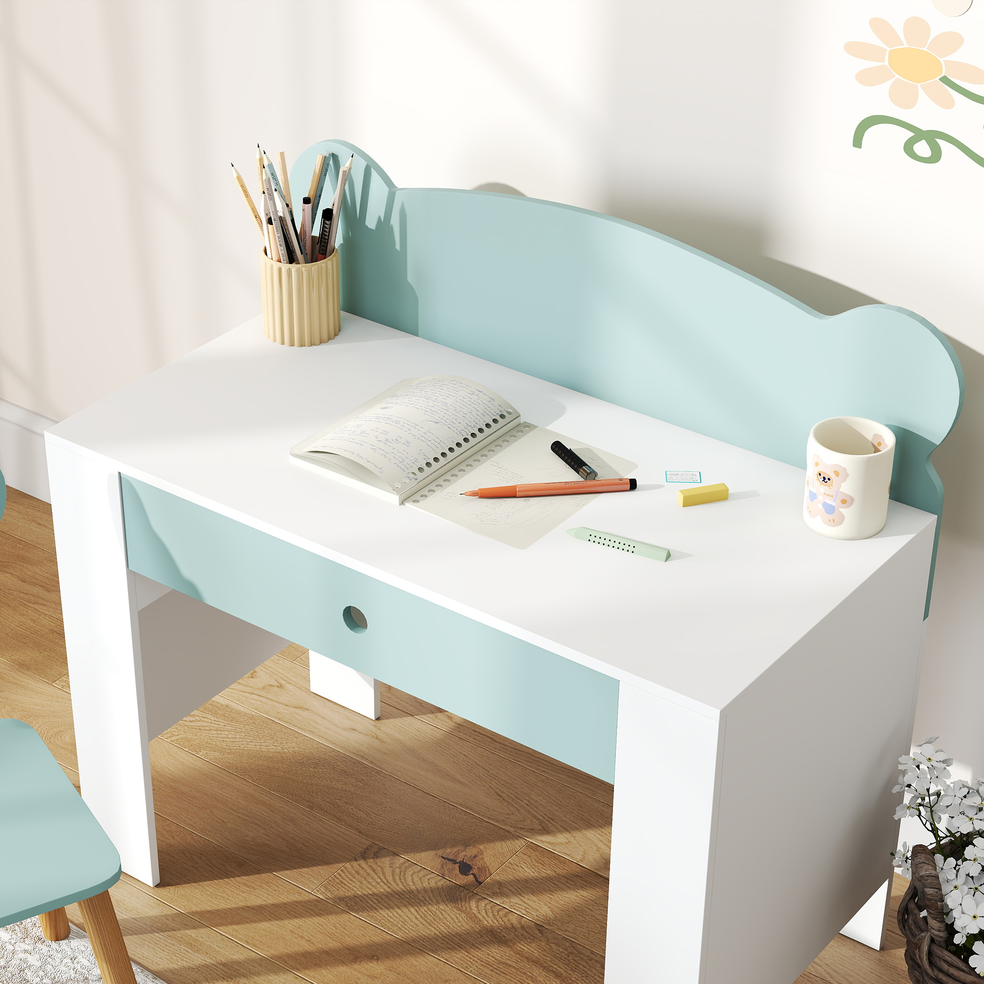 Una scrivania per bambini con cancelleria, un taccuino e una tazza con un orsetto. Una decorazione floreale sulla destra.