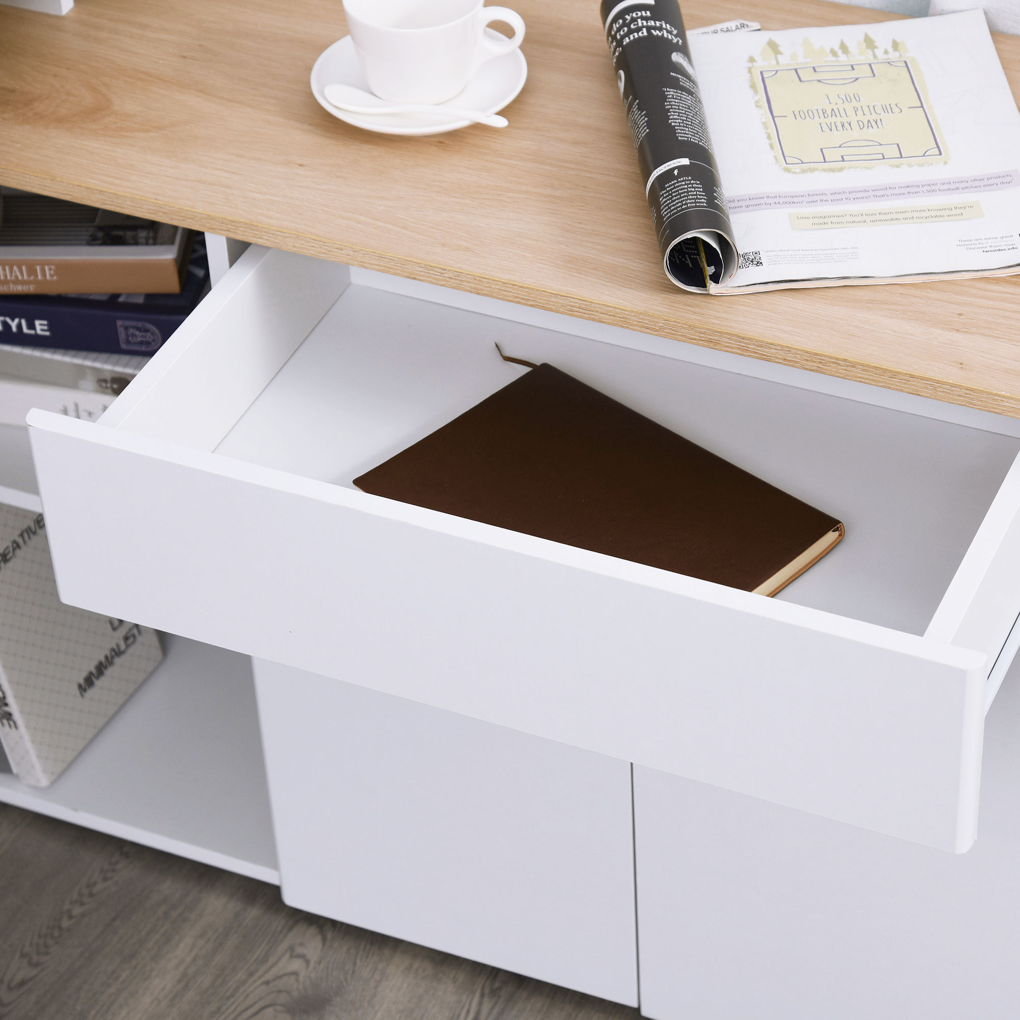 Una scrivania in legno con una rivista, una tazza e un cassetto aperto contenente un taccuino marrone. Libri visibili sotto.