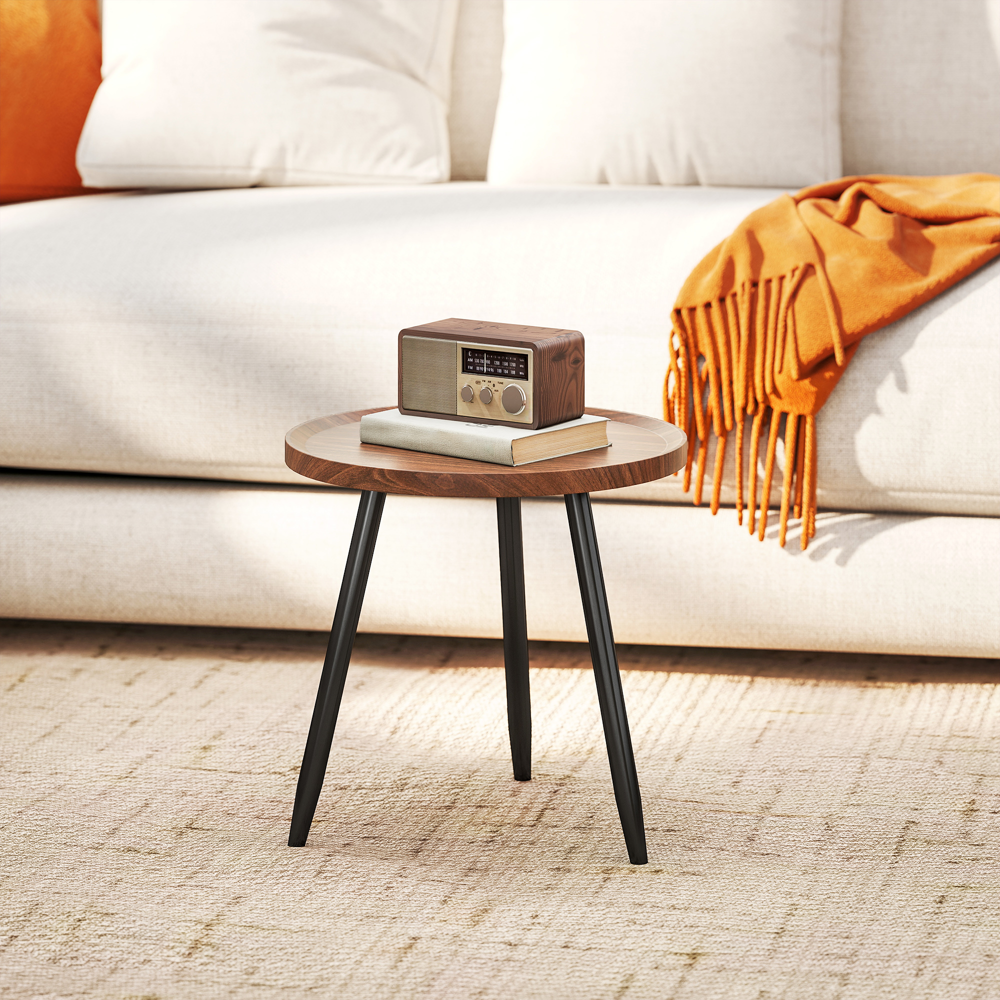 Un tavolino in legno con radio, libro e coperta arancione su tappeto beige. Un divano bianco sullo sfondo.
