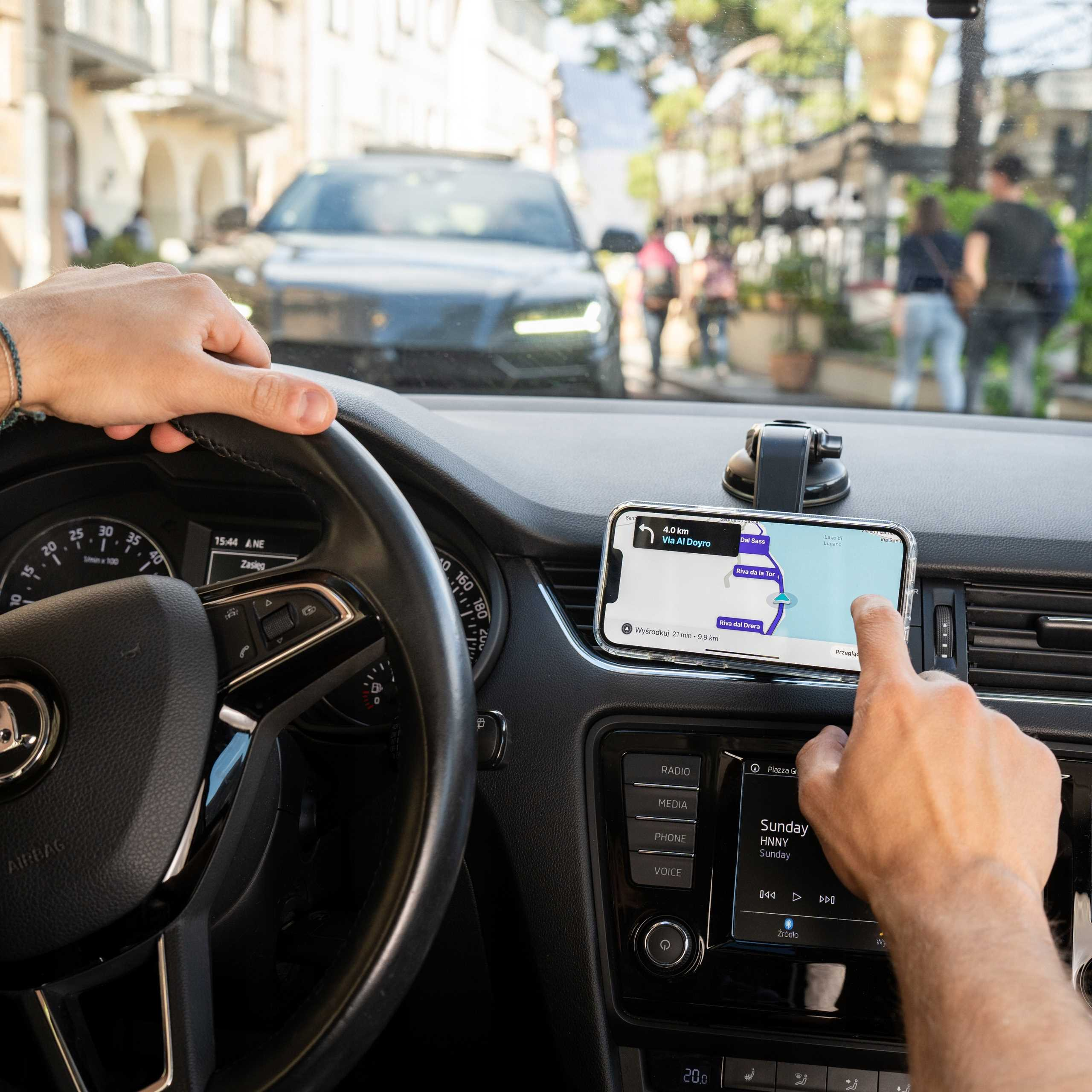Wewnątrz samochodu ręka dotyka telefonu z mapą. Widoczna kierownica. Na zewnątrz widoczne samochody i ludzie.