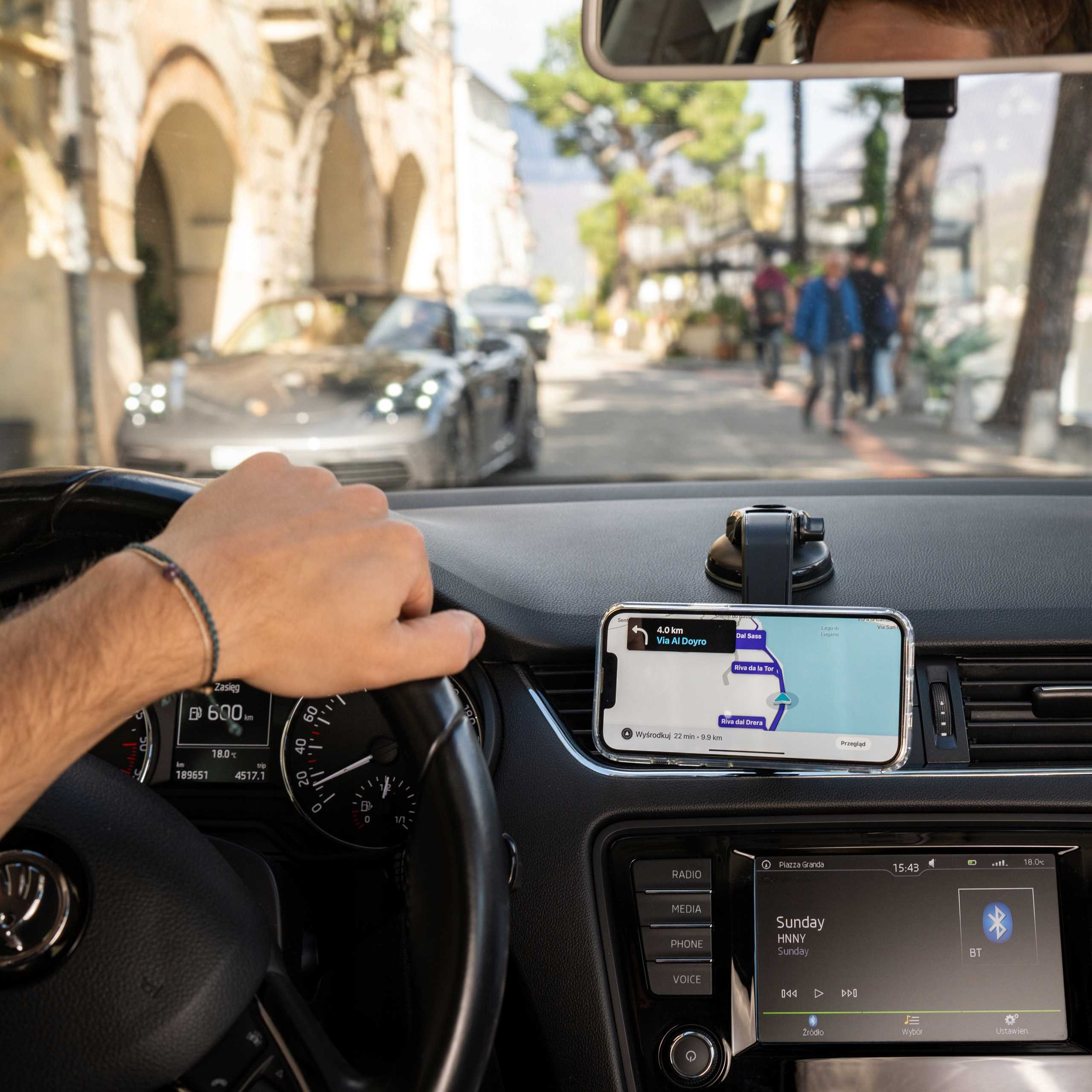 Widok kierowcy: Ręka na kierownicy, telefon z mapą, widoczna deska rozdzielcza. Scena uliczna z rozmytymi samochodami i pieszymi.