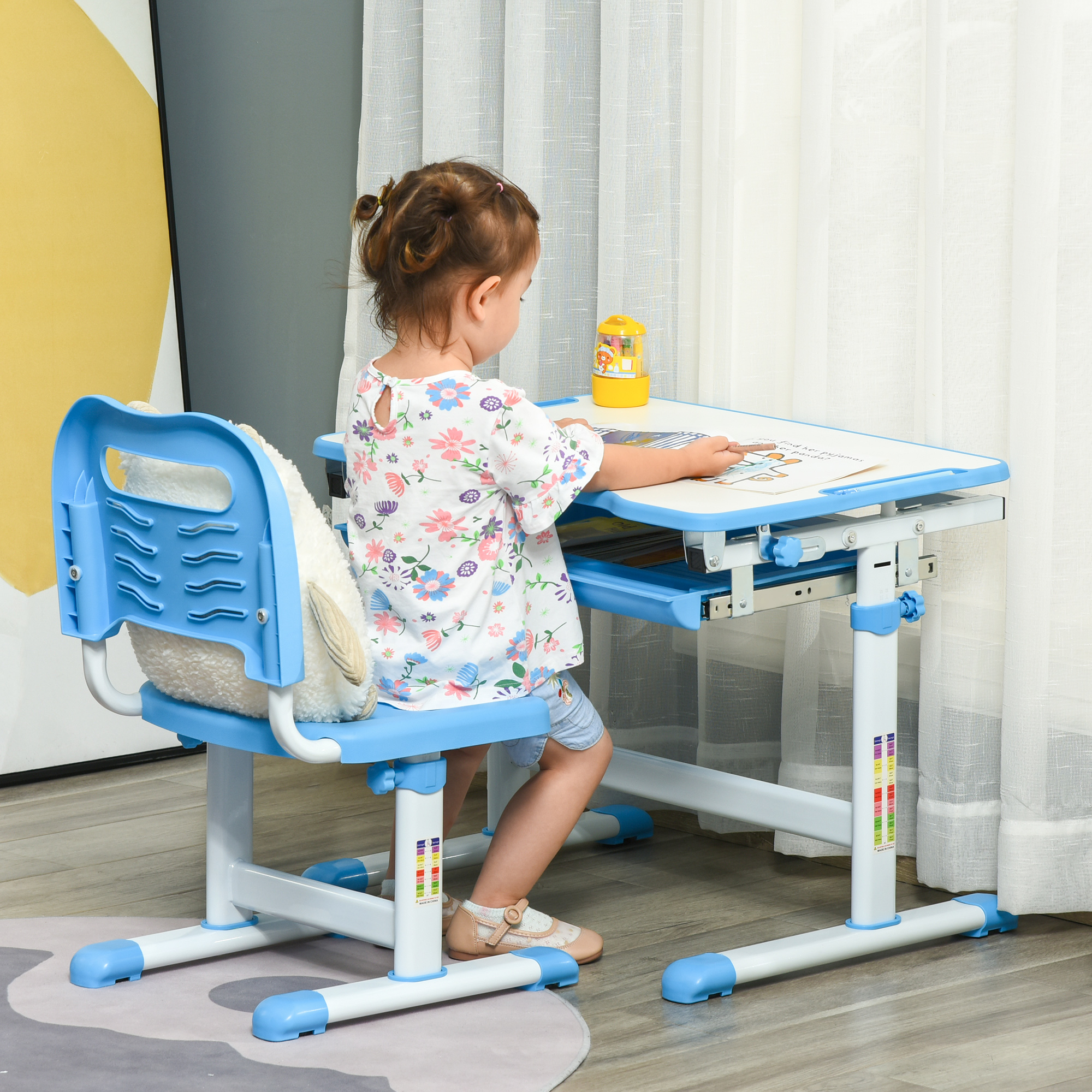 Un bambino è seduto a una scrivania blu, che guarda un libro. La scrivania è accanto a una sedia blu. Un portapenne giallo è sulla scrivania.