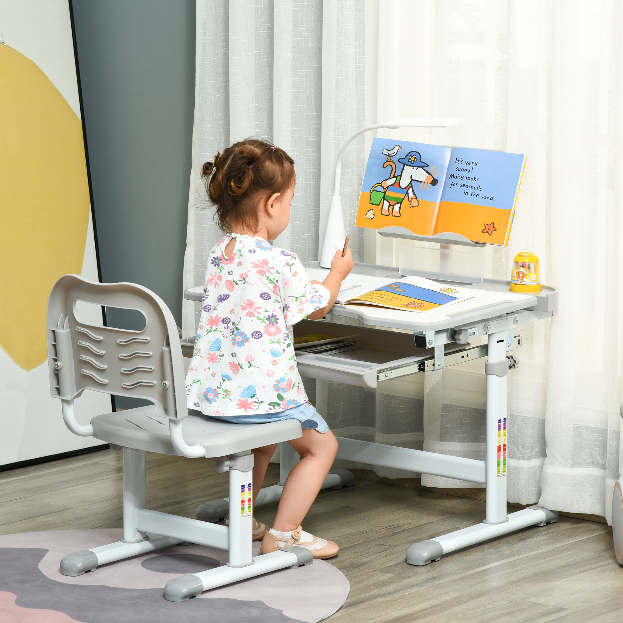 Una bambina è seduta a una scrivania grigia, leggendo un libro. Sopra il libro c'è una lampada. Lo sfondo mostra una tenda e un muro.