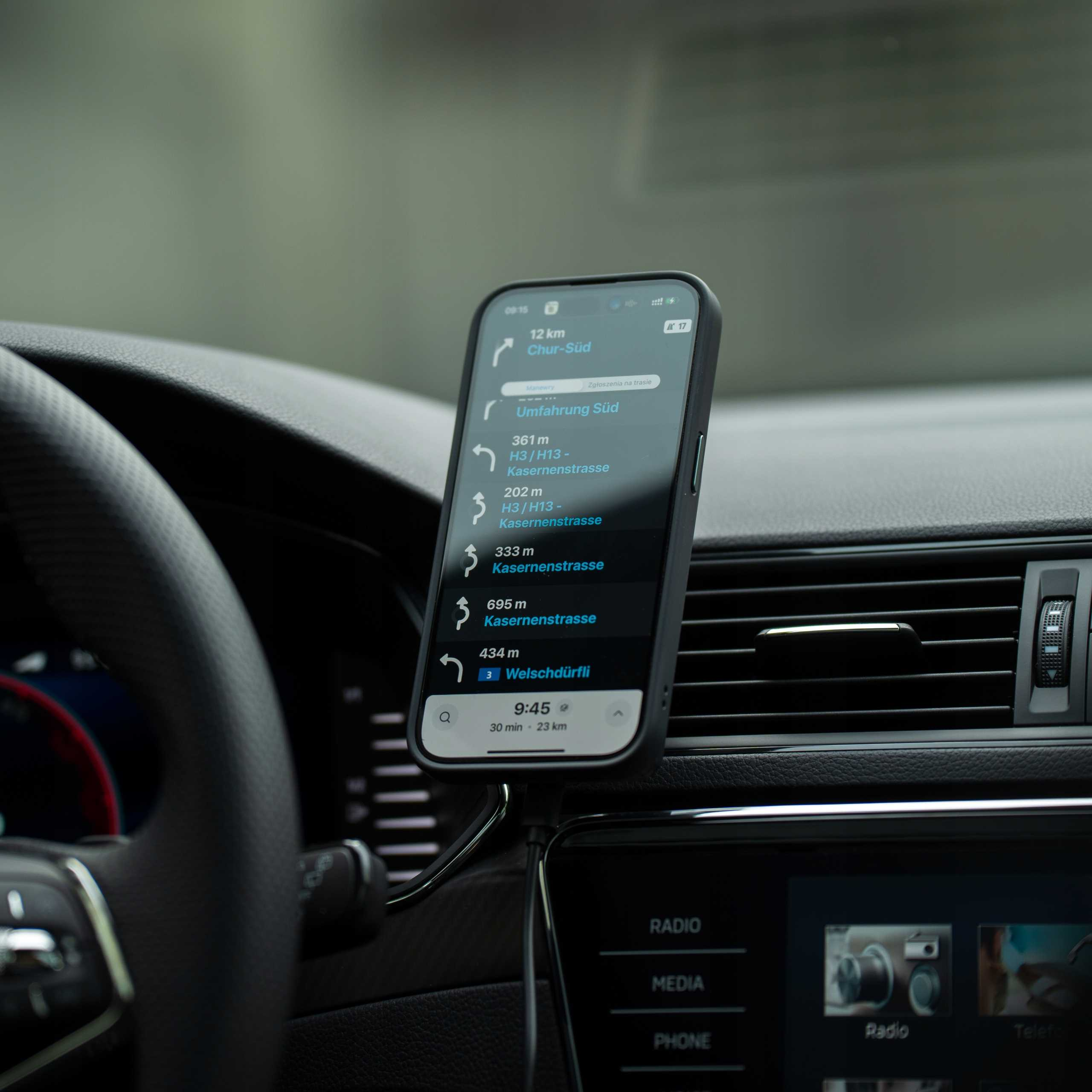 Telefon z aplikacją nawigacyjną jest zamontowany w samochodzie. Ekran pokazuje wskazówki. Widoczna kierownica i deska rozdzielcza.