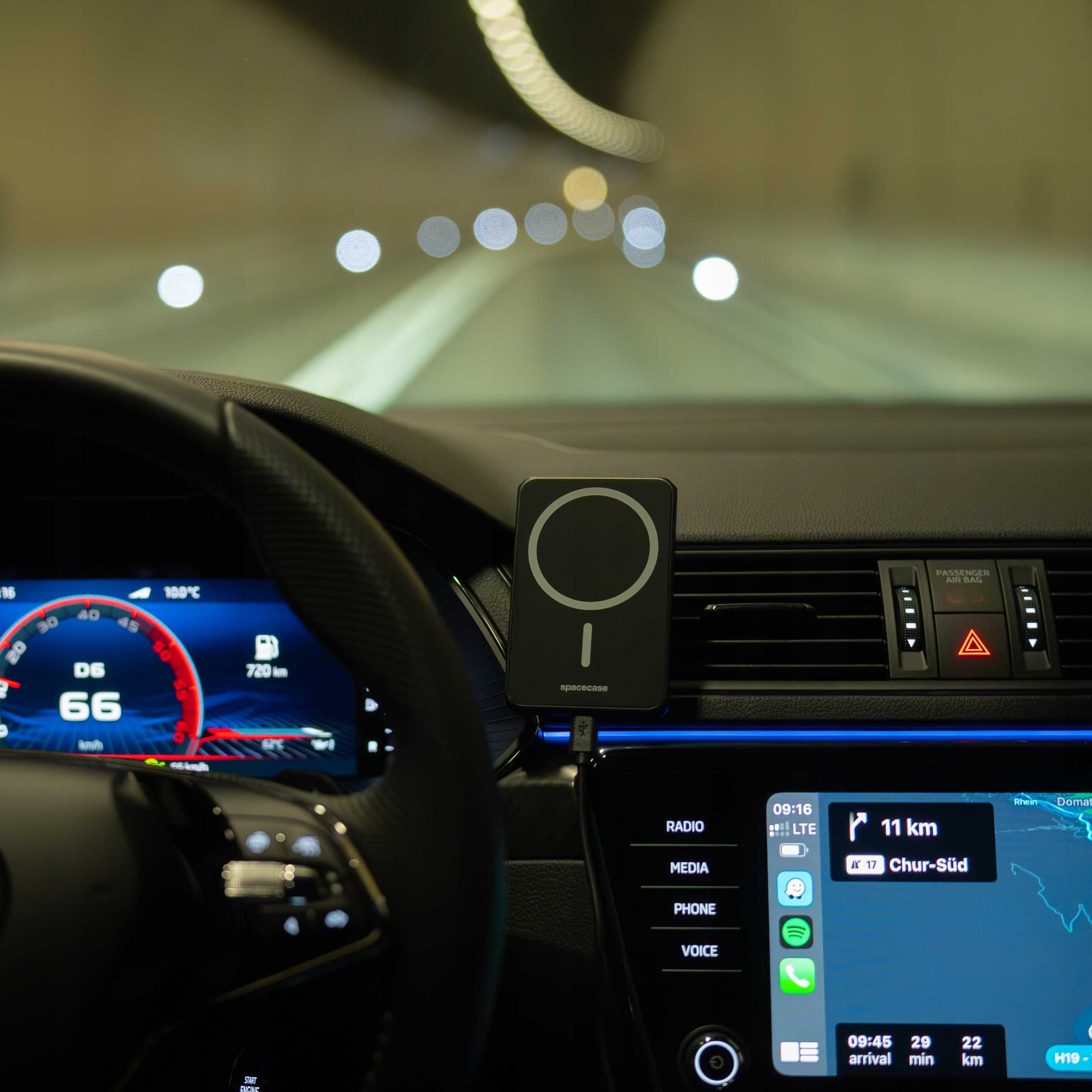 Wnętrze samochodu z deską rozdzielczą, uchwytem na telefon, wyświetlaczem i tunelem w tle. Widoczny prędkościomierz.