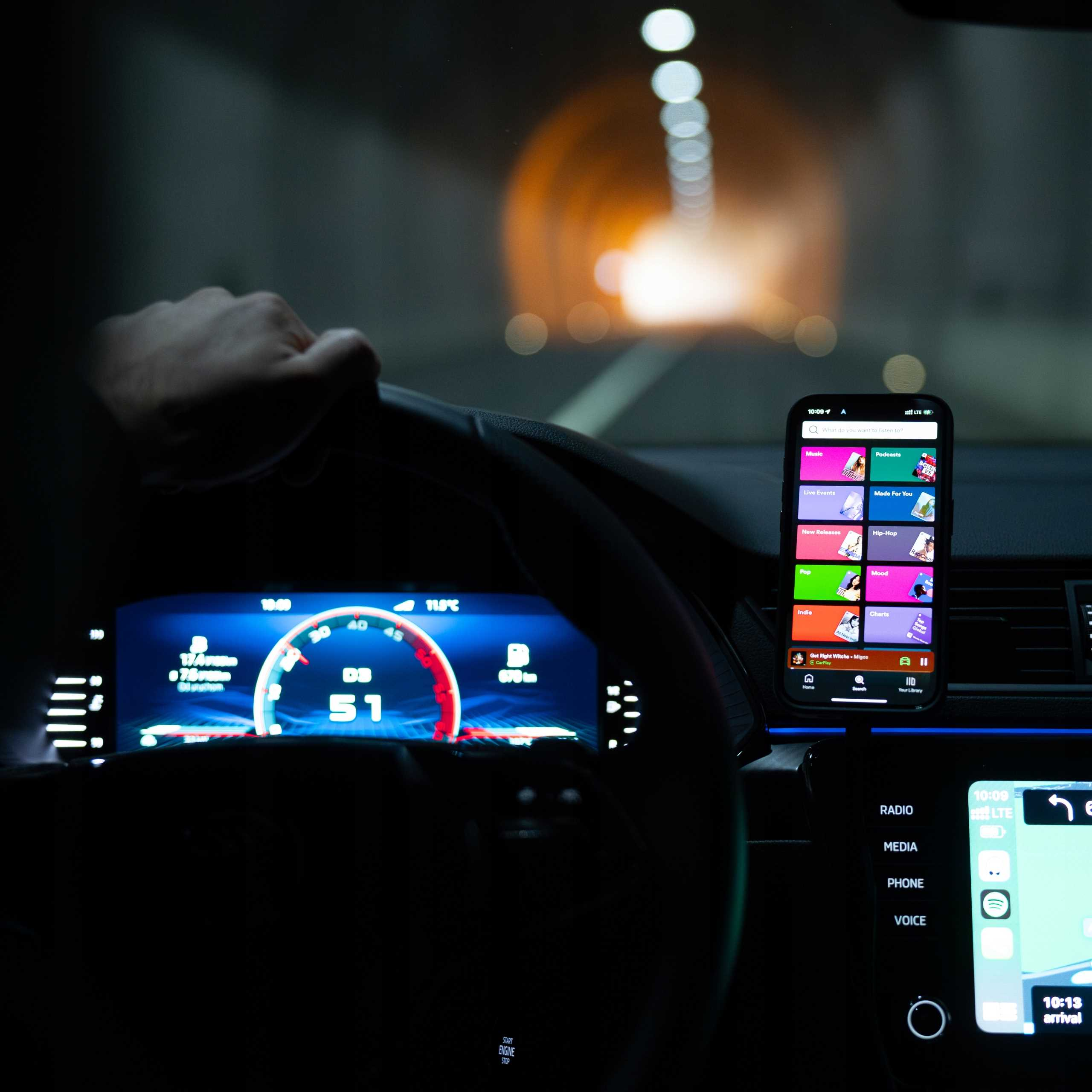 Osoba prowadzi samochód przez tunel. Telefon wyświetla aplikację muzyczną. Deska rozdzielcza pokazuje prędkość i dystans.
