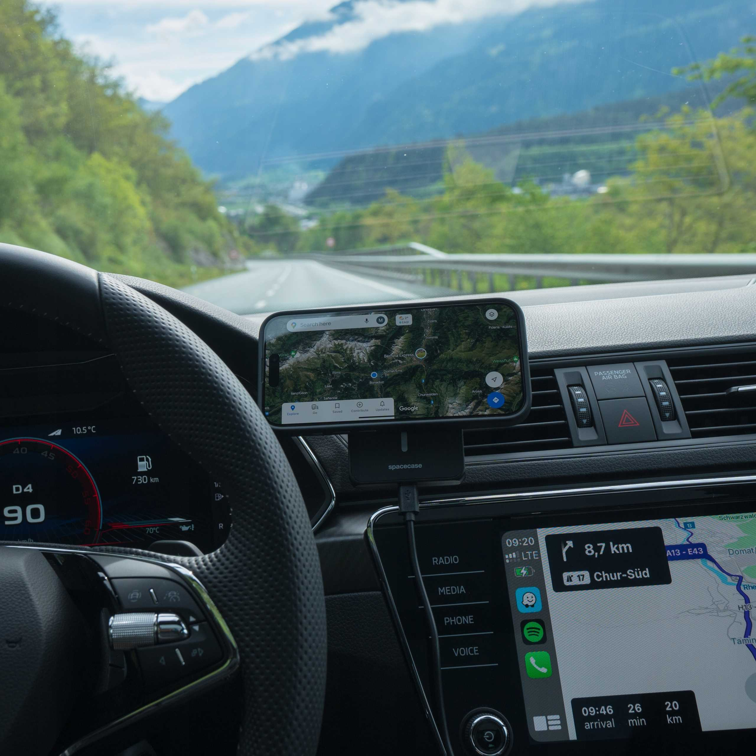 Wewnątrz samochodu, telefon z mapą na uchwycie. W tle góry. Widoczna deska rozdzielcza z prędkością i paliwem.