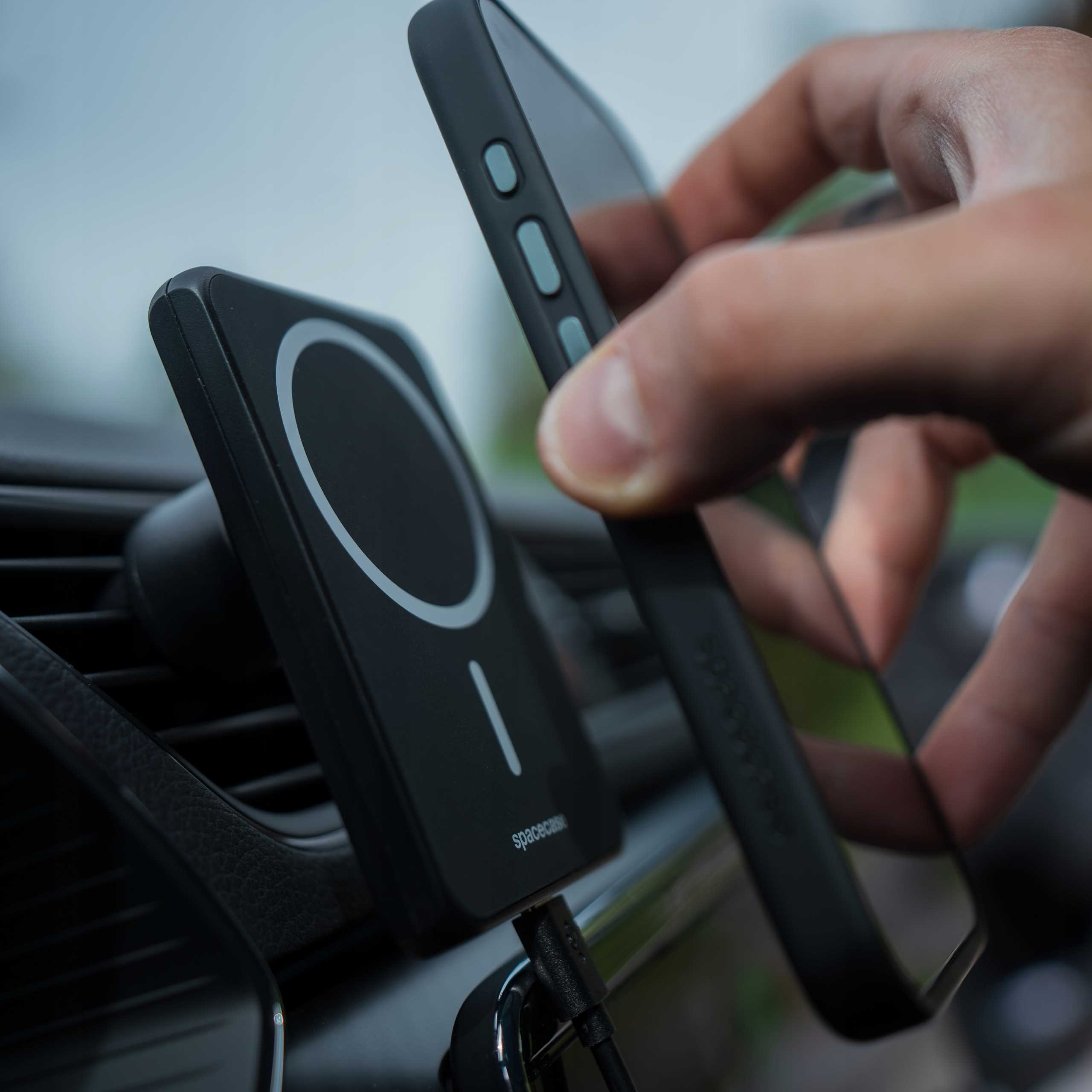 Dłoń trzymająca telefon w pobliżu czarnej ładowarki samochodowej. Ładowarka jest zamontowana na otworze wentylacyjnym. Telefon ma czarne etui.