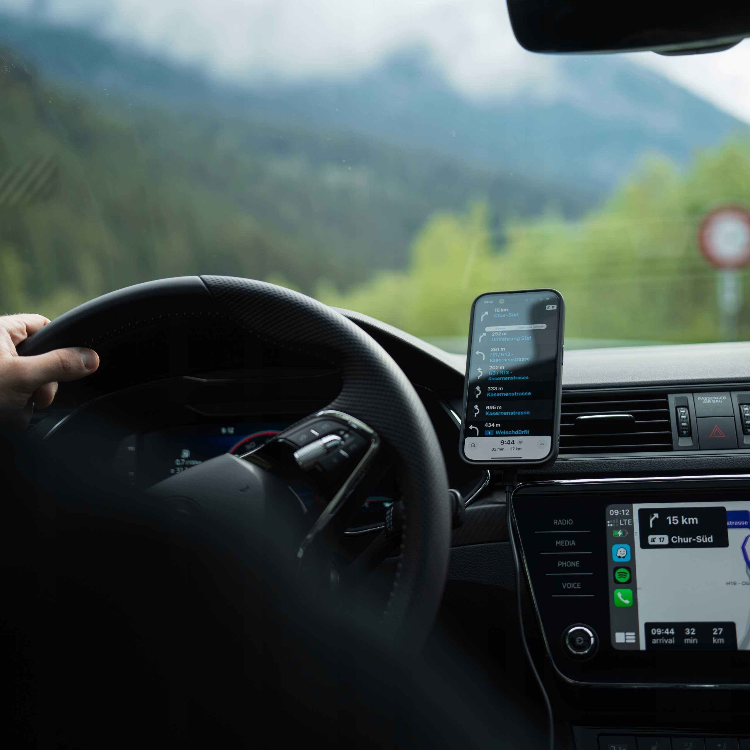 Wnętrze samochodu, ręka na kierownicy. Telefon z aplikacją nawigacyjną. Na zewnątrz widoczne góry i droga.
