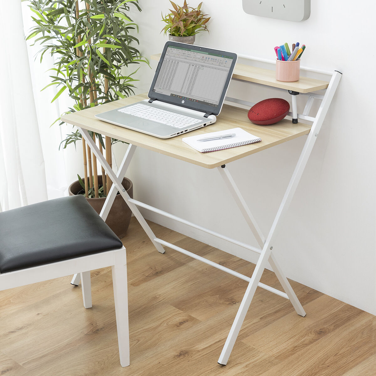 Un portátil sobre un escritorio con una silla delante. Plantas en el fondo. Un reloj en la pared.