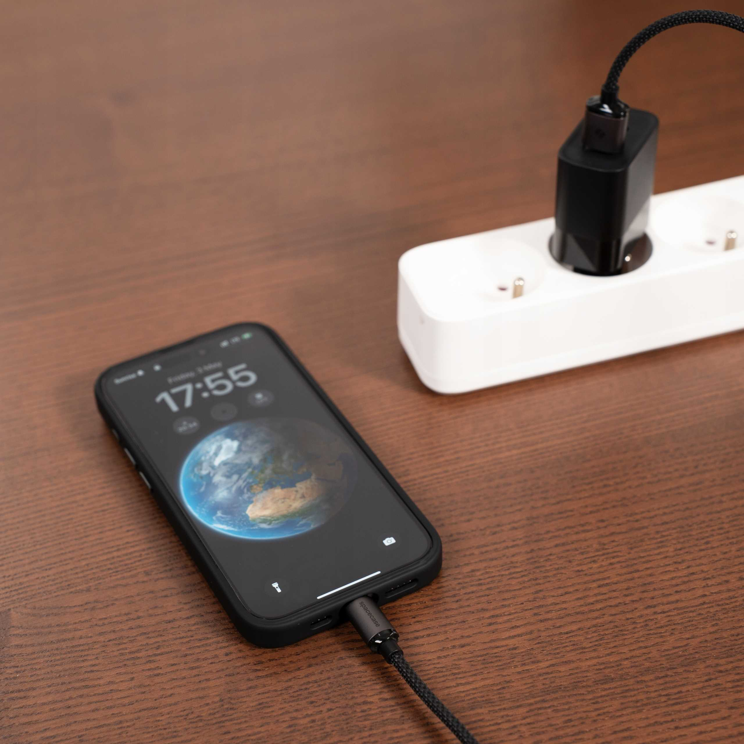 Telefon ładujący się na drewnianej powierzchni. Ładowarka jest podłączona do gniazdka. Telefon wyświetla godzinę 17:55.