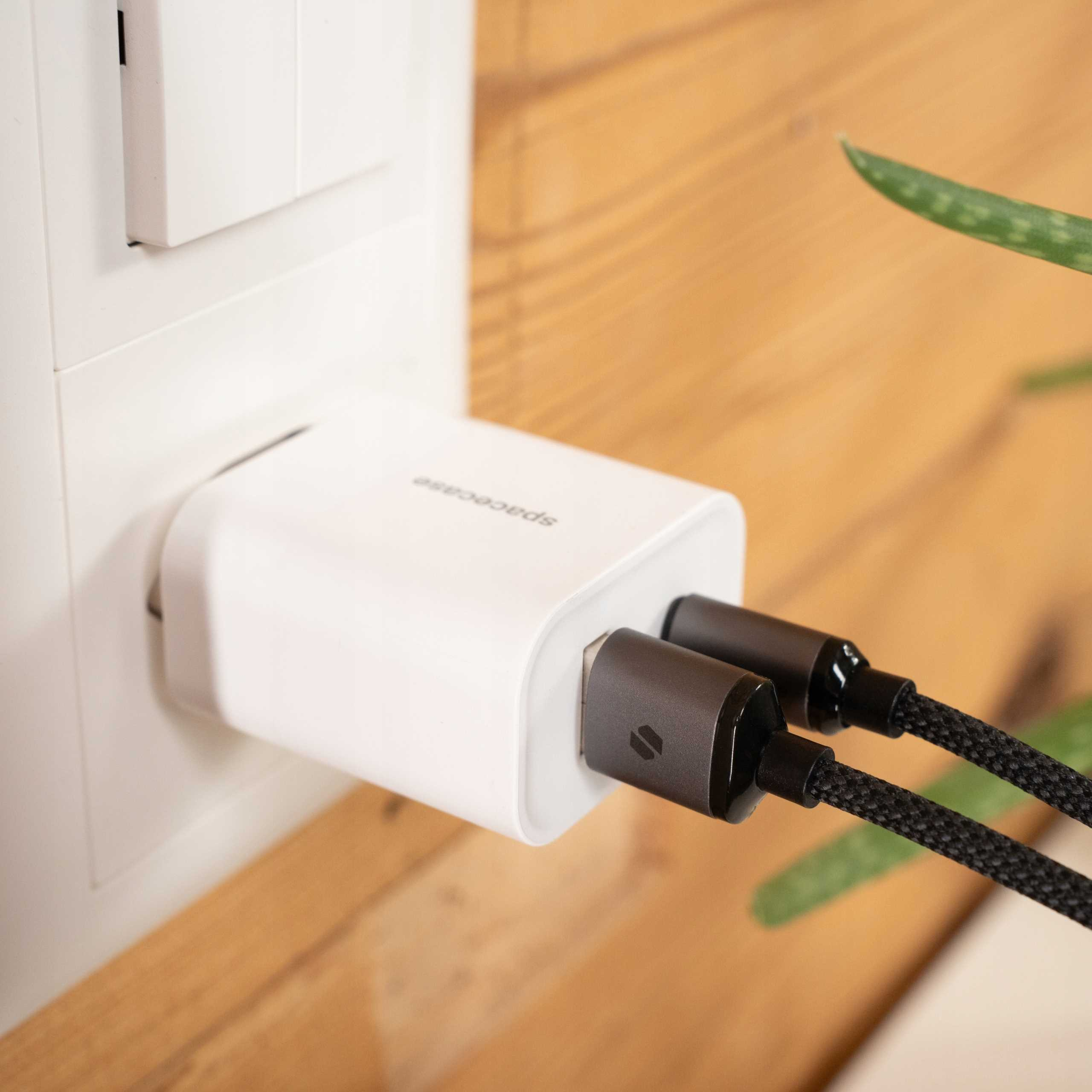 Biała ładowarka podłączona do gniazdka ściennego z dwoma czarnymi kablami. Tło to drewniana powierzchnia i zielona roślina.