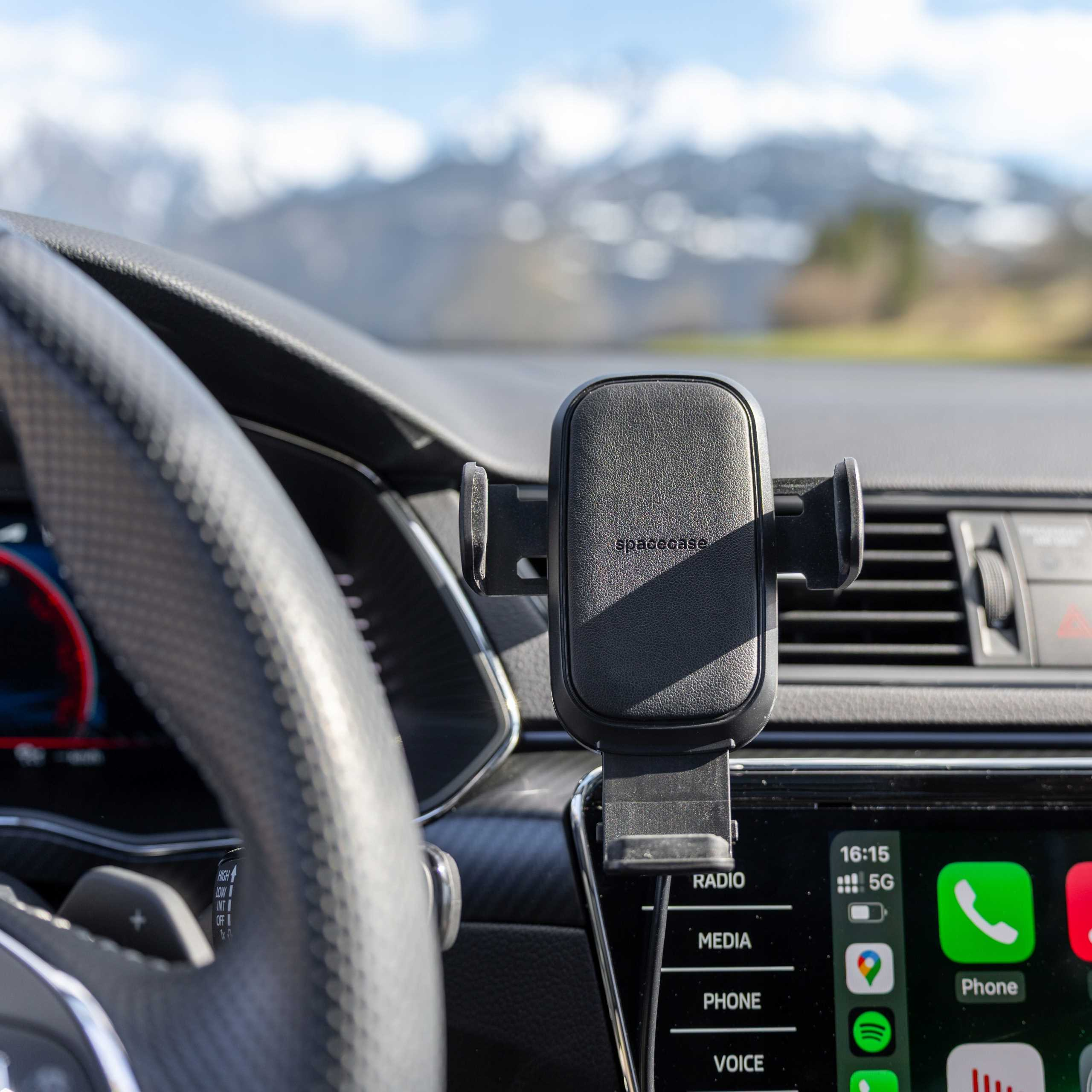 Wnętrze samochodu. Czarny uchwyt na telefon z napisem 'spacecase'. Kierownica po lewej, ekran informacyjno-rozrywkowy po prawej, widoczne góry.