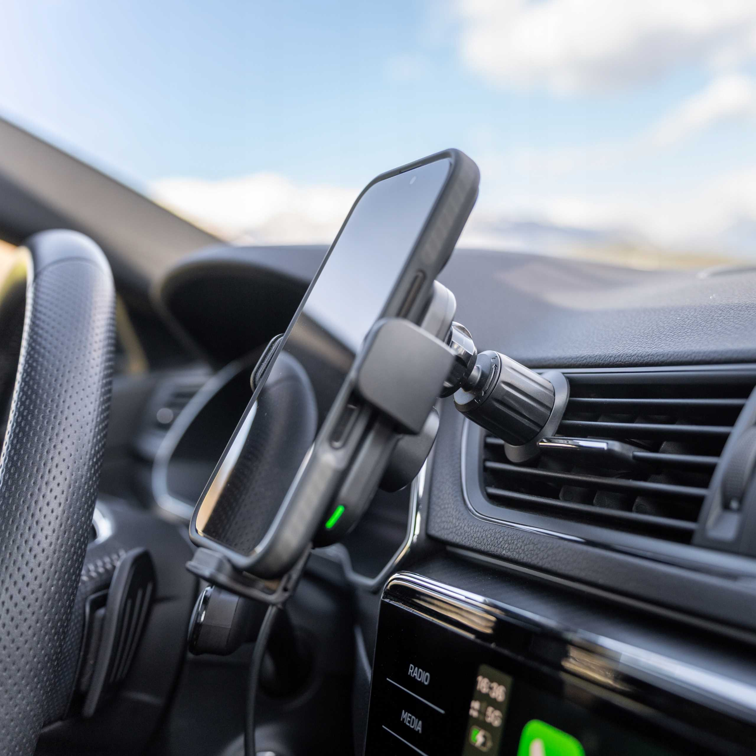 Telefon jest zamontowany w samochodzie. Telefon jest czarny. Kierownica widoczna po lewej. Niebo jest niebieskie.