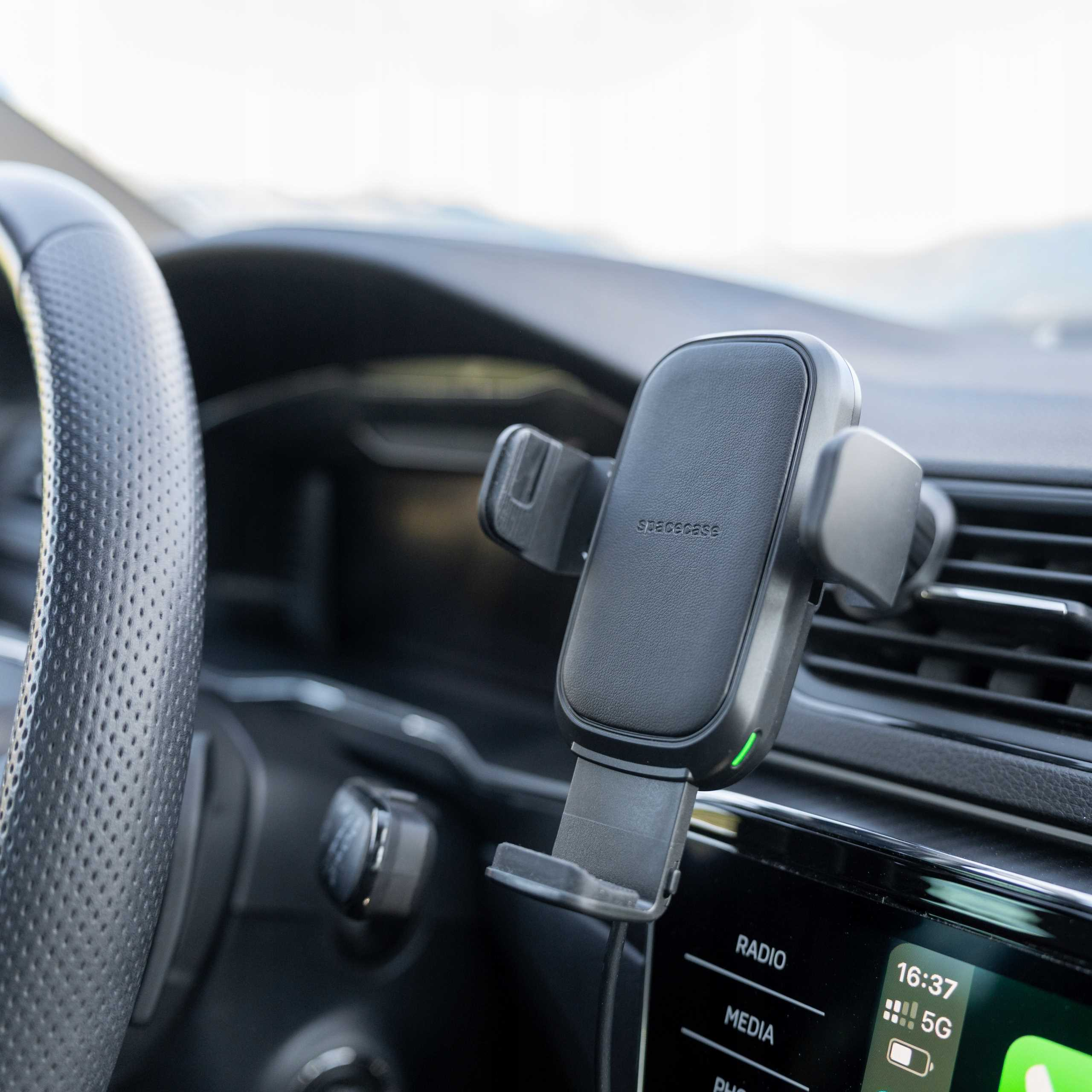 Czarny uchwyt na telefon samochodowy zamontowany na desce rozdzielczej. Widoczna kierownica. Ekran pokazuje 16:37 i 5G.