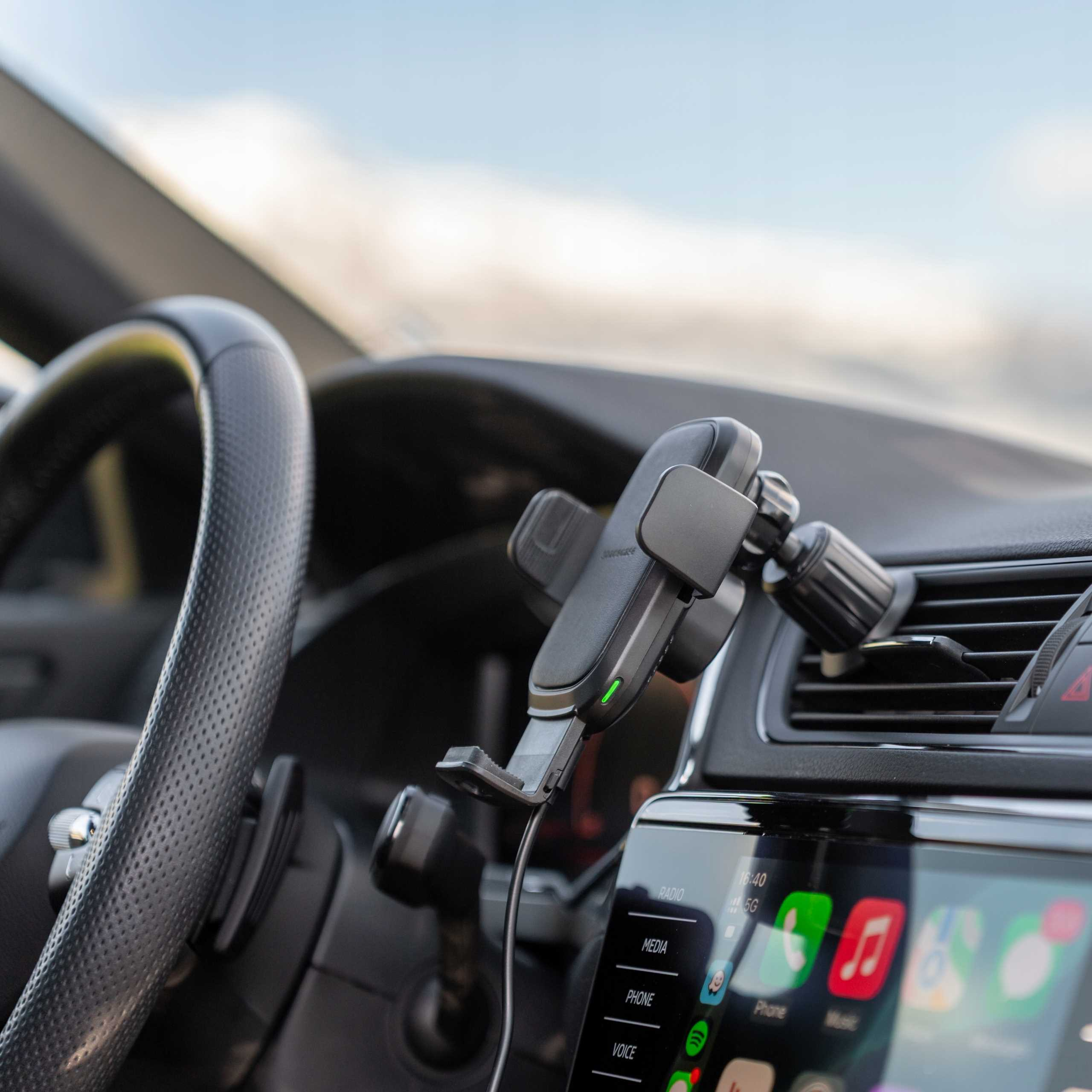 Widok wnętrza samochodu z kierownicą, uchwytem na telefon i ekranem informacyjnym. Widoczne niebieskie niebo.