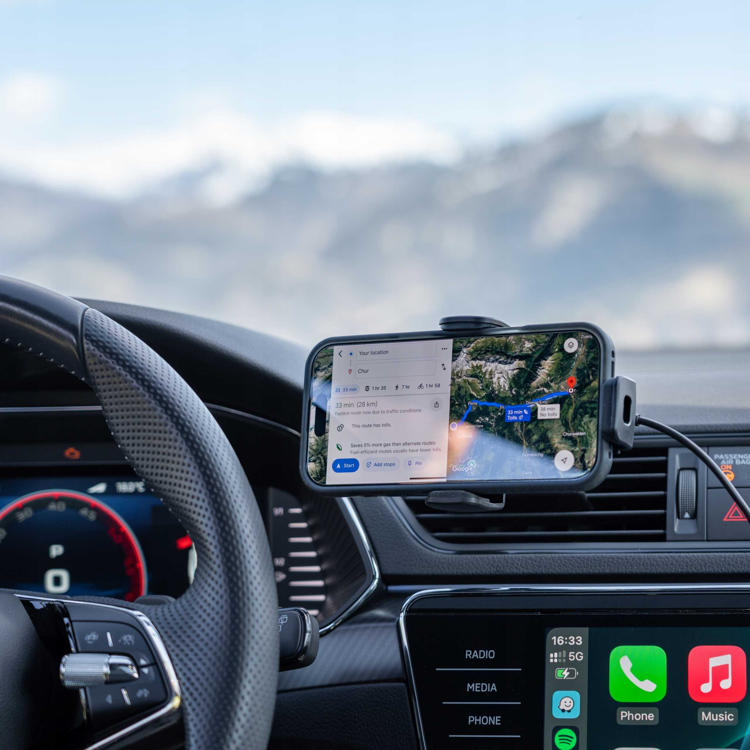 Wnętrze samochodu z telefonem wyświetlającym aplikację nawigacyjną. Kierownica i deska rozdzielcza widoczne, z górami w tle.