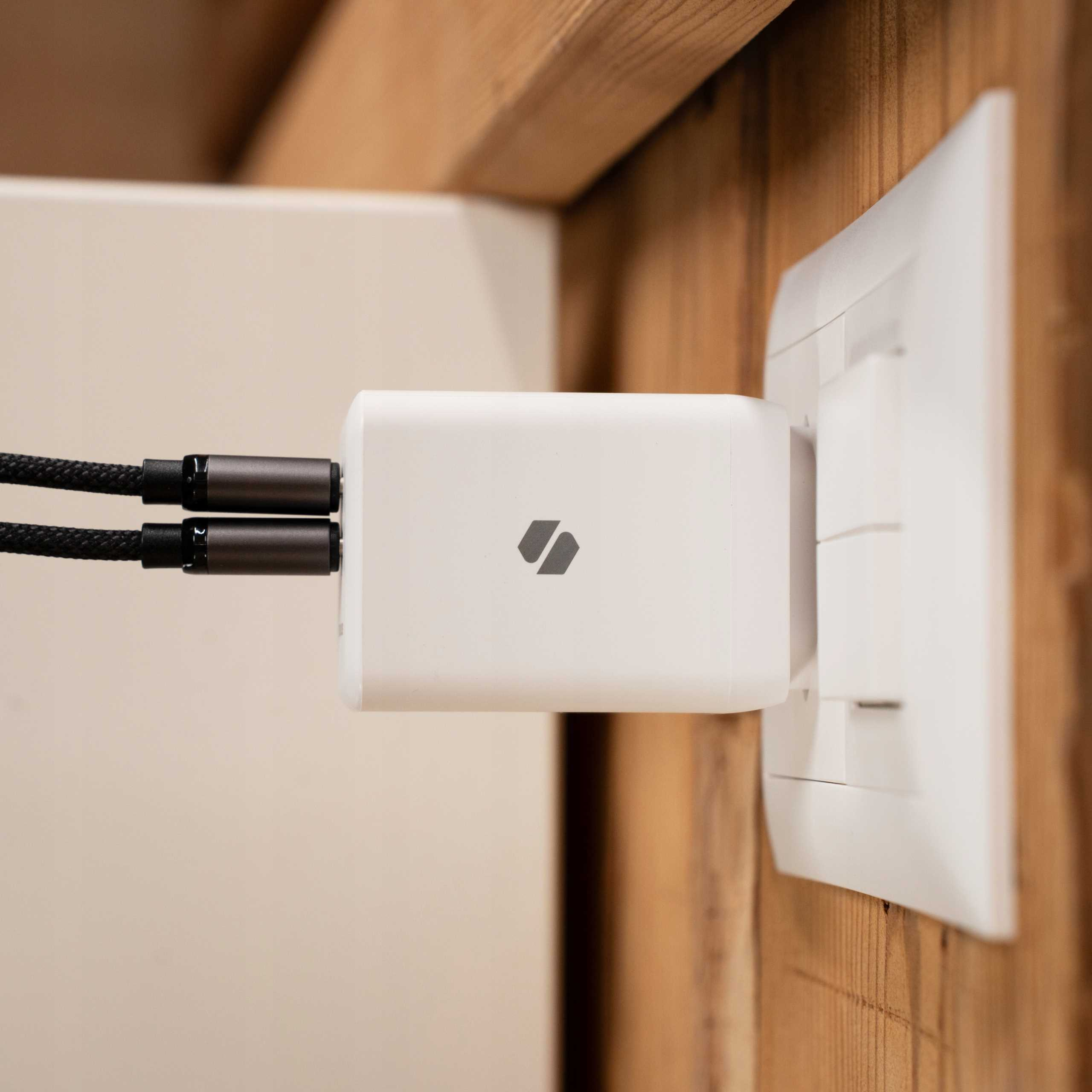 Biała ładowarka z logo jest podłączona do białego gniazdka. Czarne kable są podłączone do ładowarki. Tło jest drewniane.