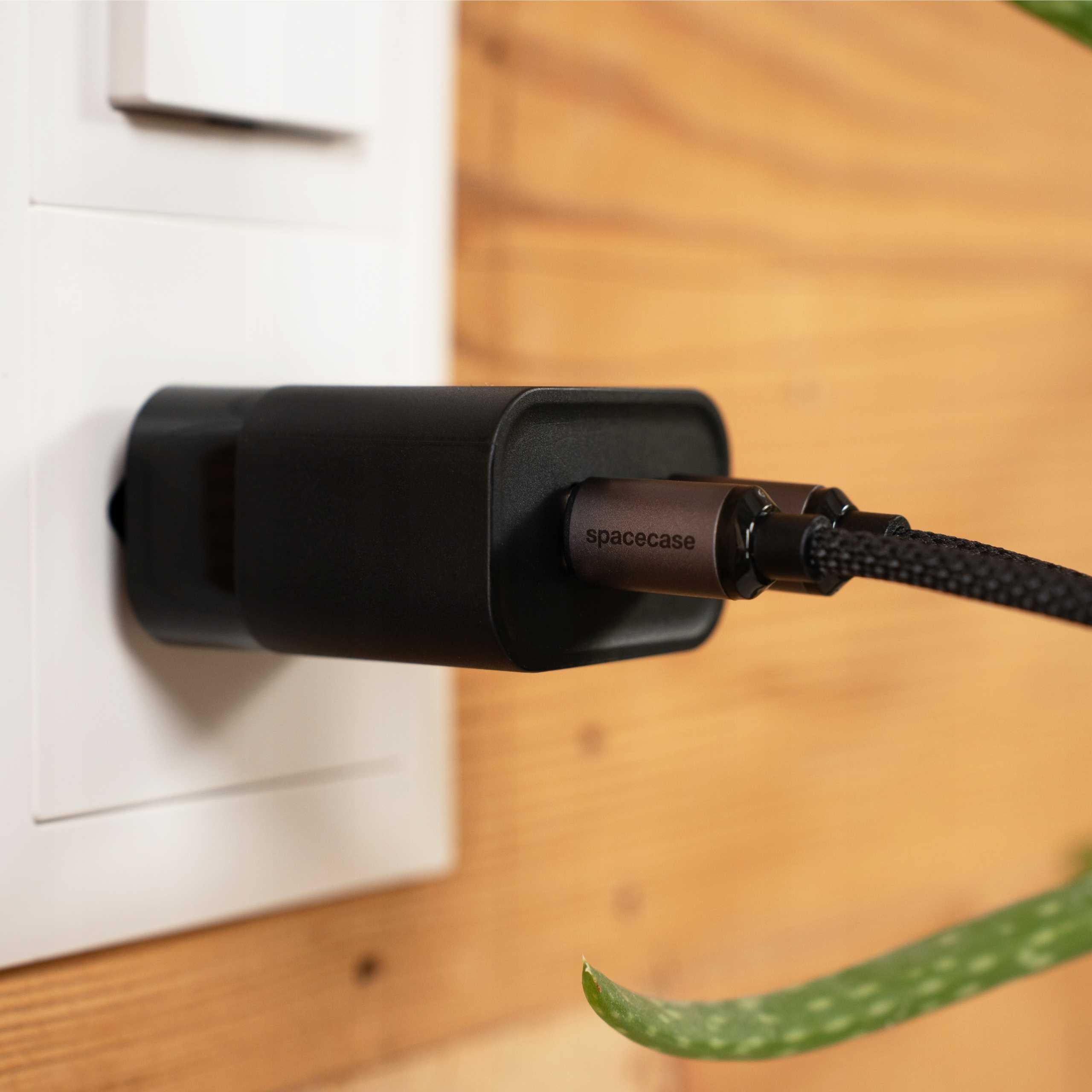 Czarna ładowarka jest podłączona do białego gniazdka. Na kablu widnieje napis 'spacecase'. Tło to drewno. Zielony liść jest w rogu.