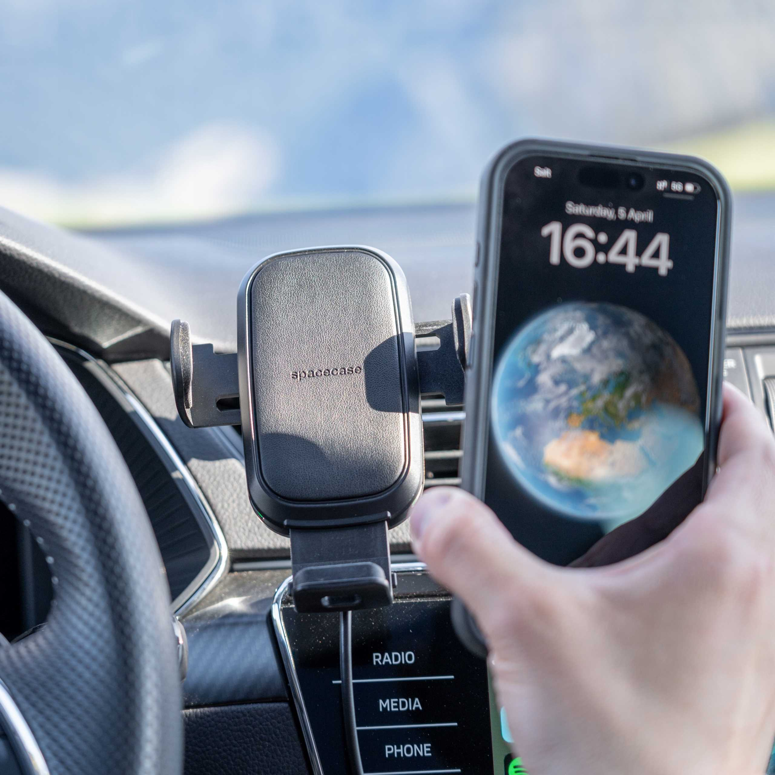 Telefon w uchwycie samochodowym wyświetla godzinę. Dłoń trzyma telefon. Uchwyt jest czarny, tło to wnętrze samochodu.