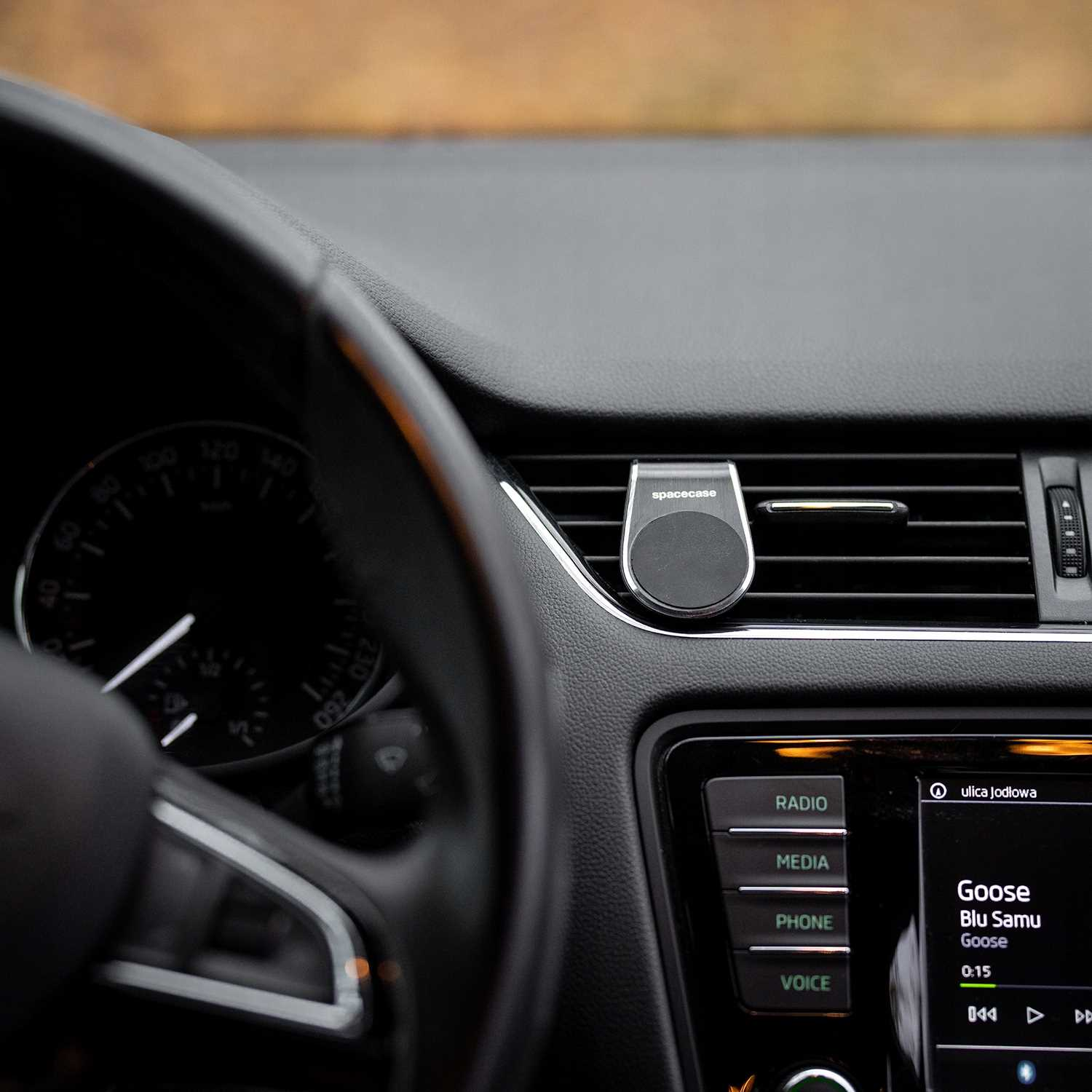 Wnętrze samochodu z kierownicą, deską rozdzielczą i uchwytem na telefon. Wyświetlacz radia pokazuje piosenkę i wykonawcę. Widoczny nawiew i uchwyt.
