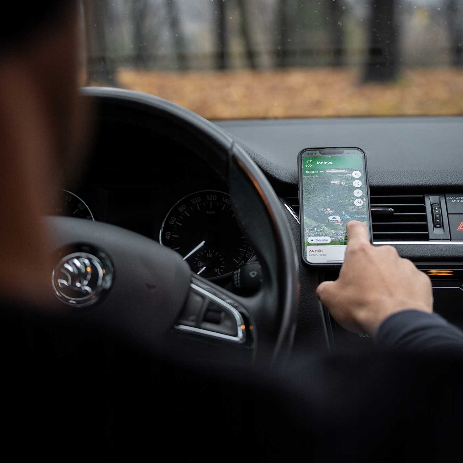 Osoba w samochodzie korzysta z telefonu wyświetlającego mapę. Telefon jest zamontowany na desce rozdzielczej. Widoczna jest kierownica.