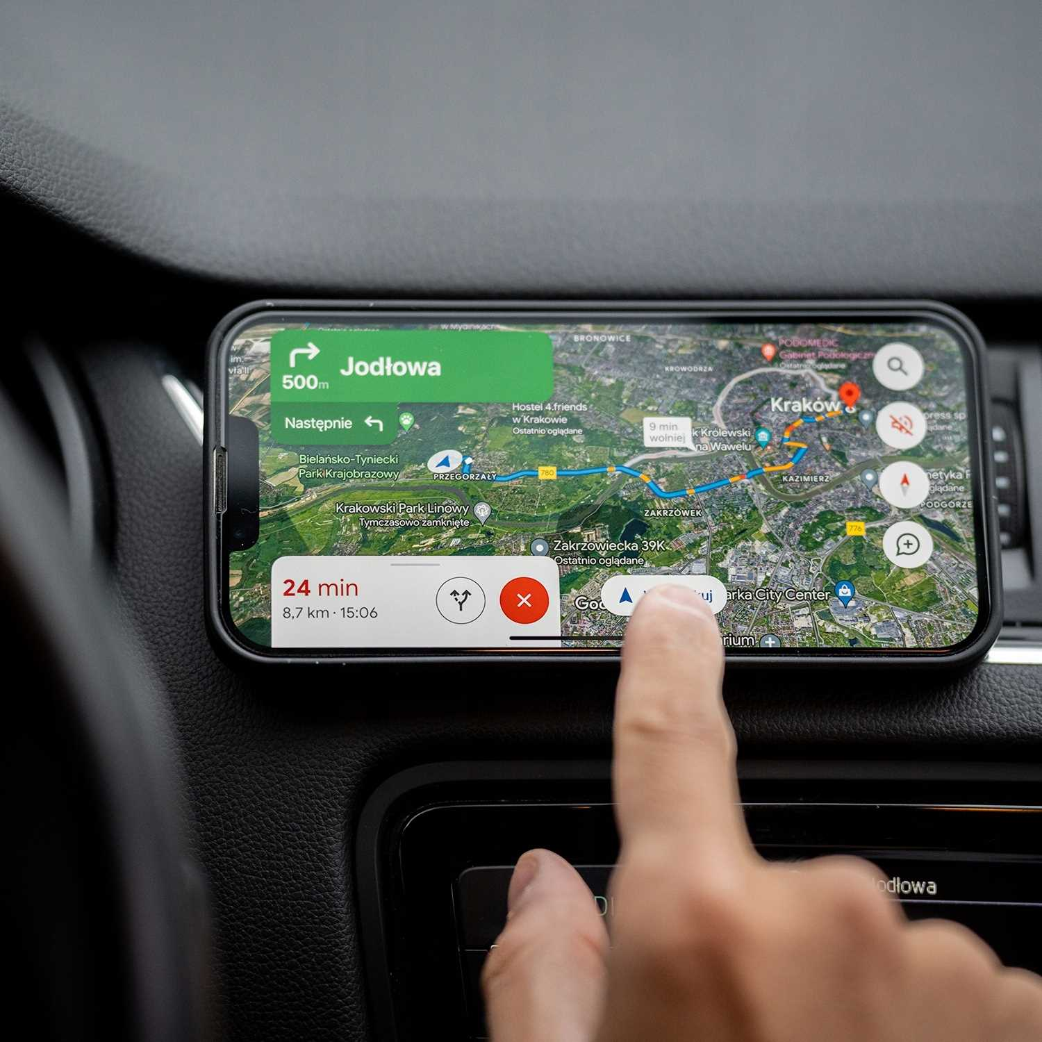 Telefon wyświetla mapę z wskazówkami. Palec dotyka ekranu. Deska rozdzielcza samochodu jest widoczna.
