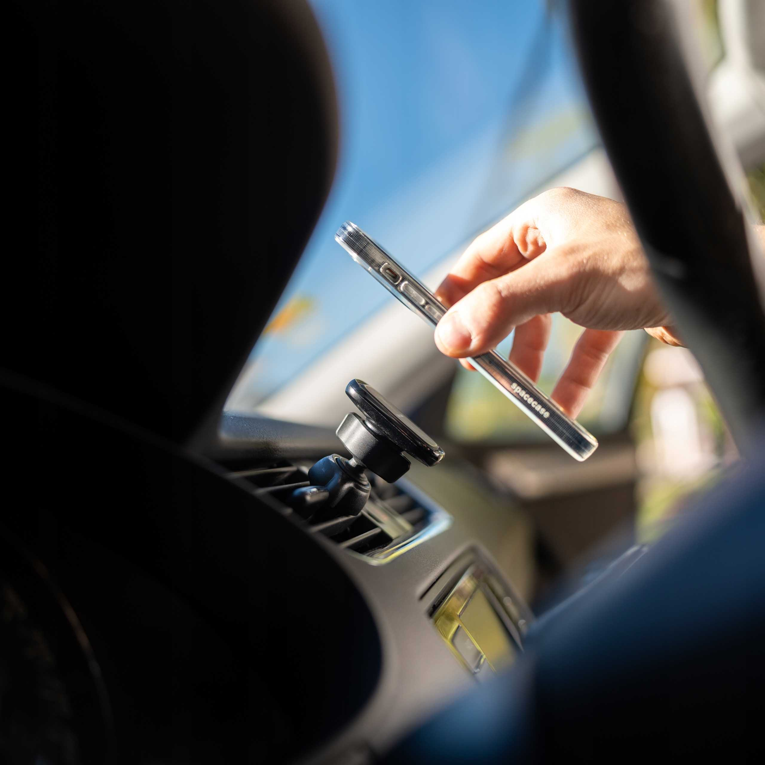 Dłoń trzyma przezroczyste etui na telefon nad uchwytem wentylacyjnym. Niebieskie niebo widoczne przez przednią szybę.