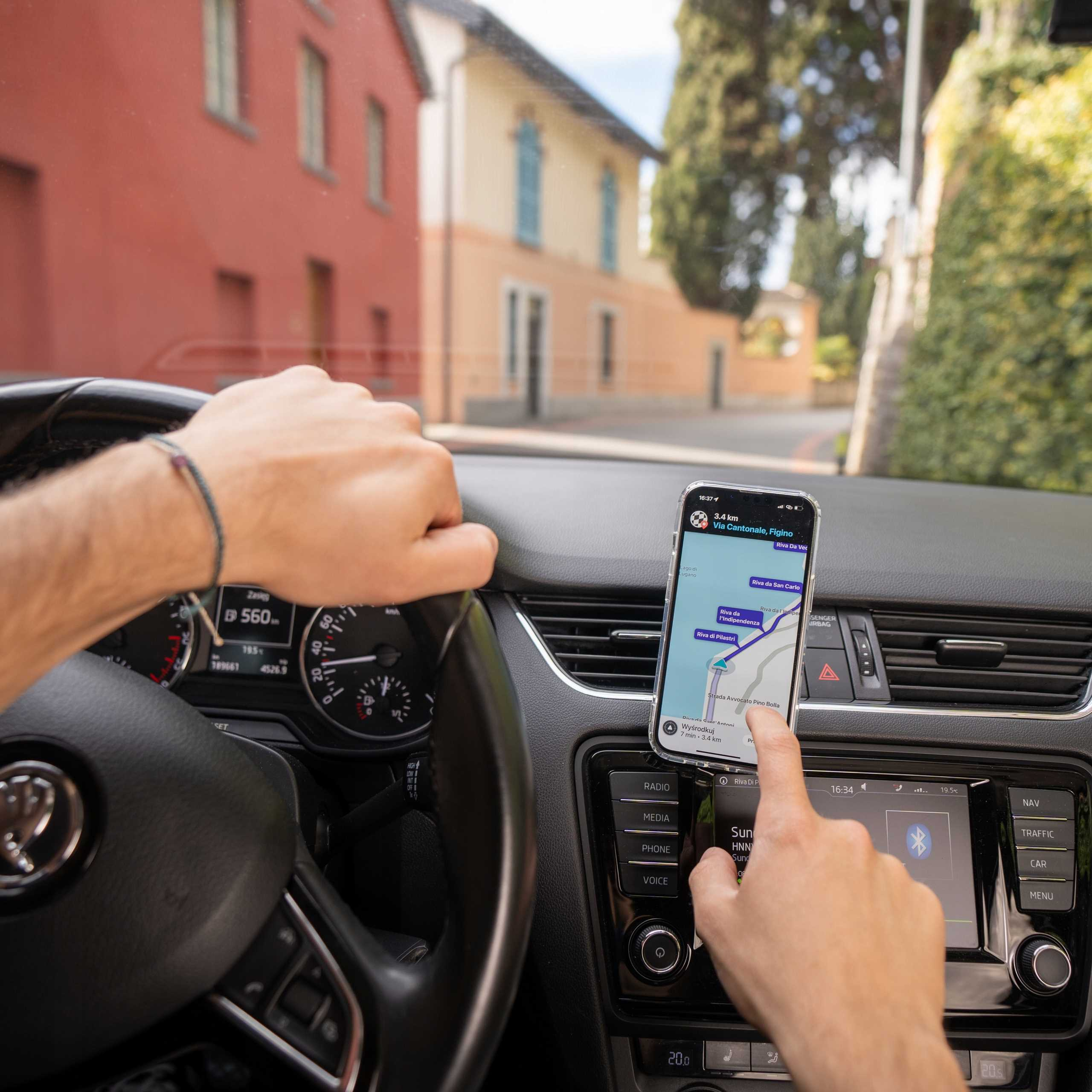 Ręce na kierownicy i telefon z mapą. Budynki i drzewa na zewnątrz.