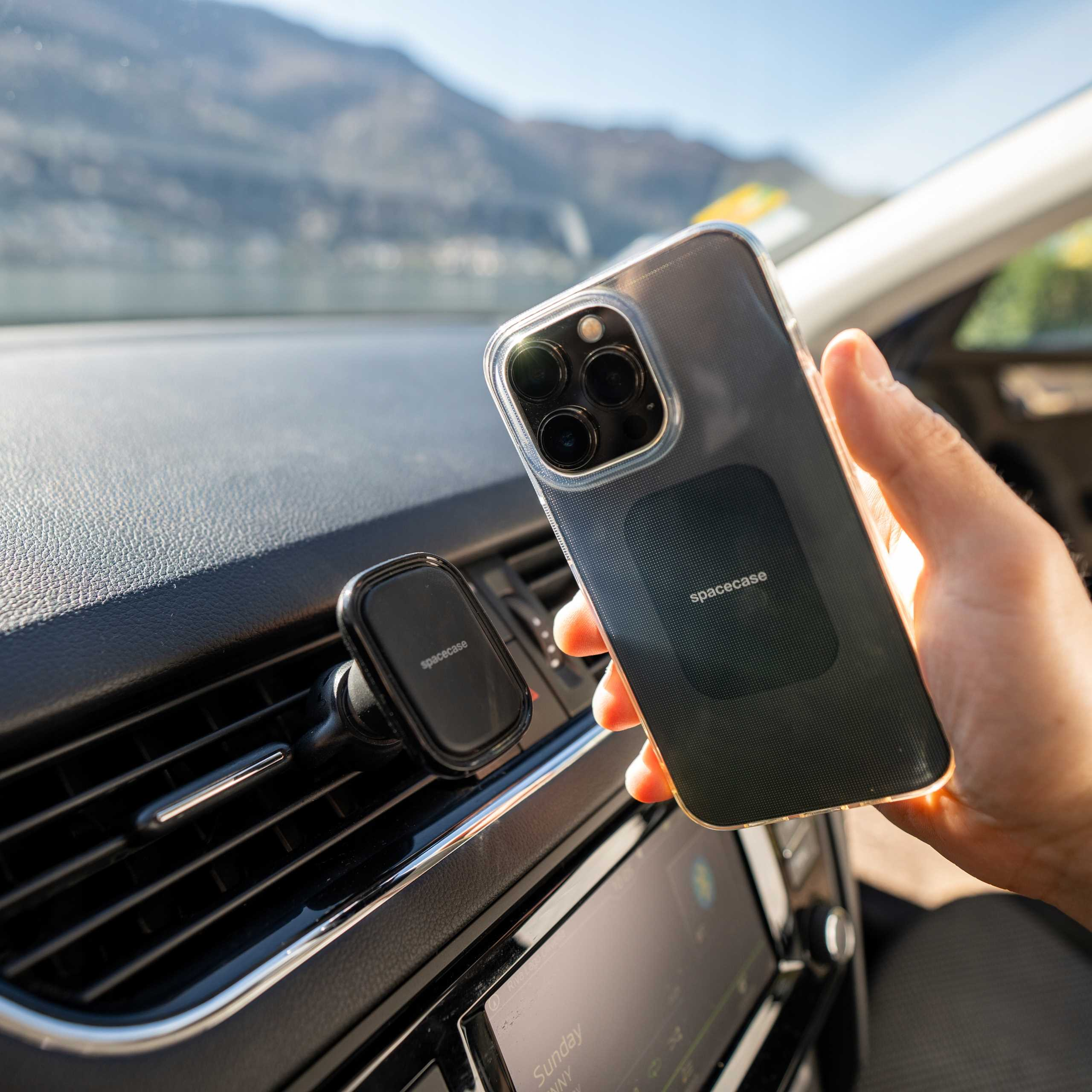 Ręka trzyma telefon w etui. Uchwyt samochodowy na telefon jest na desce rozdzielczej. Przez przednią szybę widać góry.