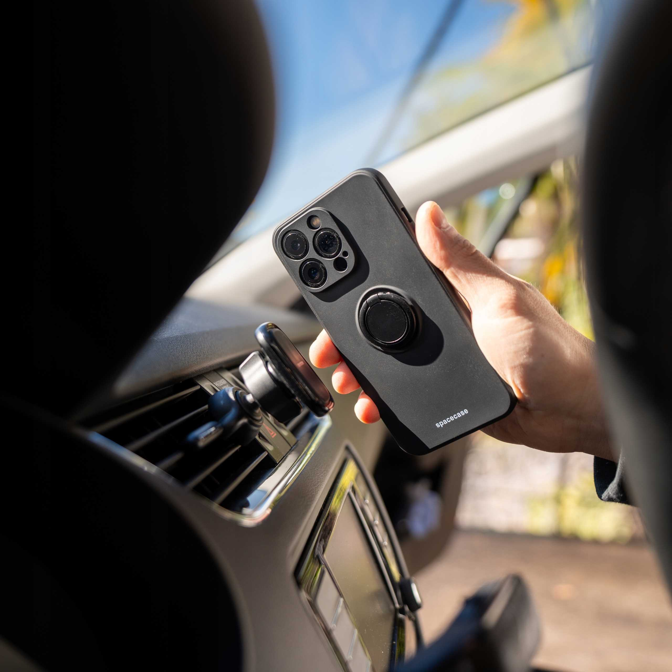 Dłoń trzymająca czarny telefon z uchwytem na pierścień w samochodzie. Widoczny jest magnetyczny uchwyt na telefon. Tło pokazuje wnętrze samochodu.