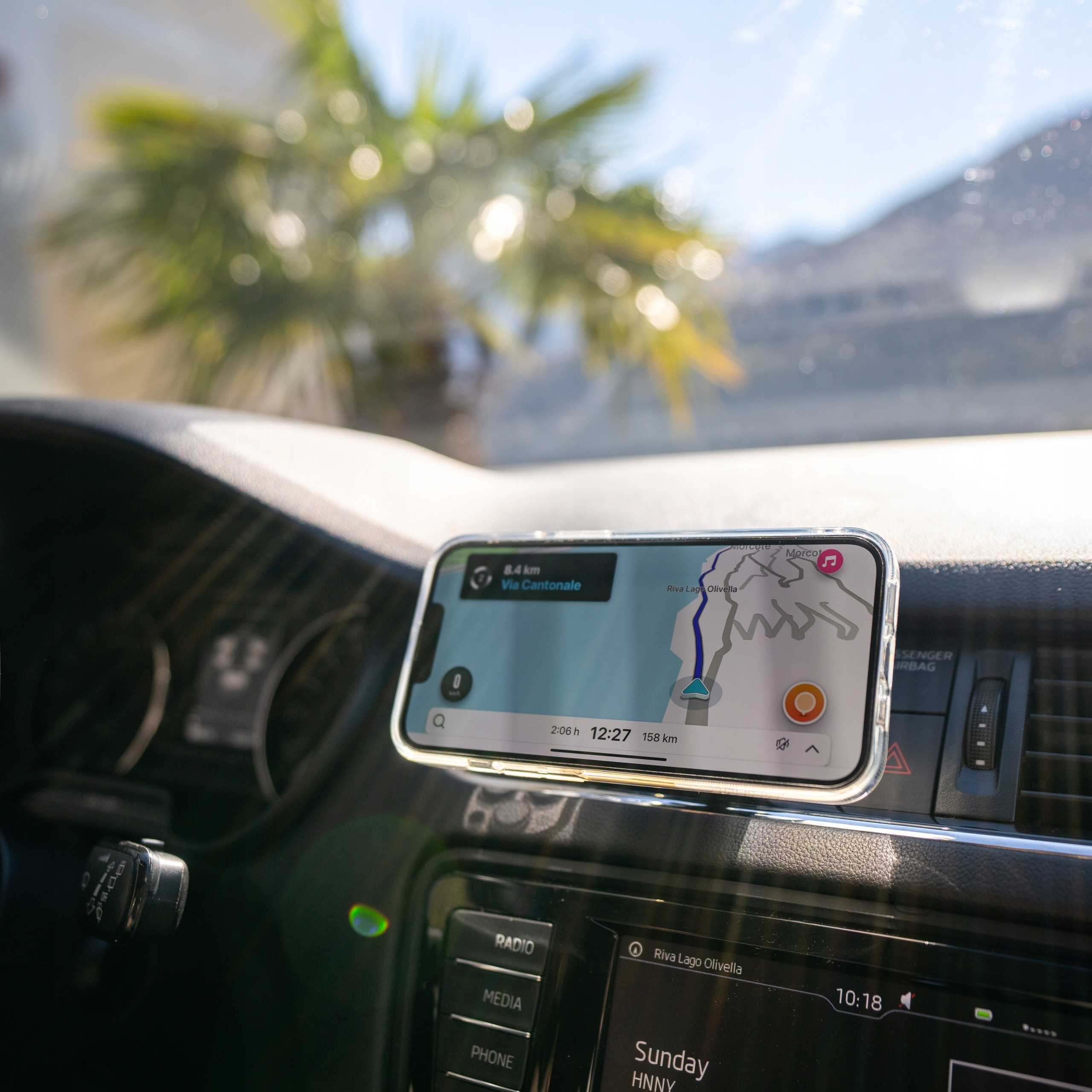 Widok na deskę rozdzielczą z telefonem wyświetlającym mapę. Drzewo palmowe widoczne przez przednią szybę. Godzina 12:27. Dystans 158 km.