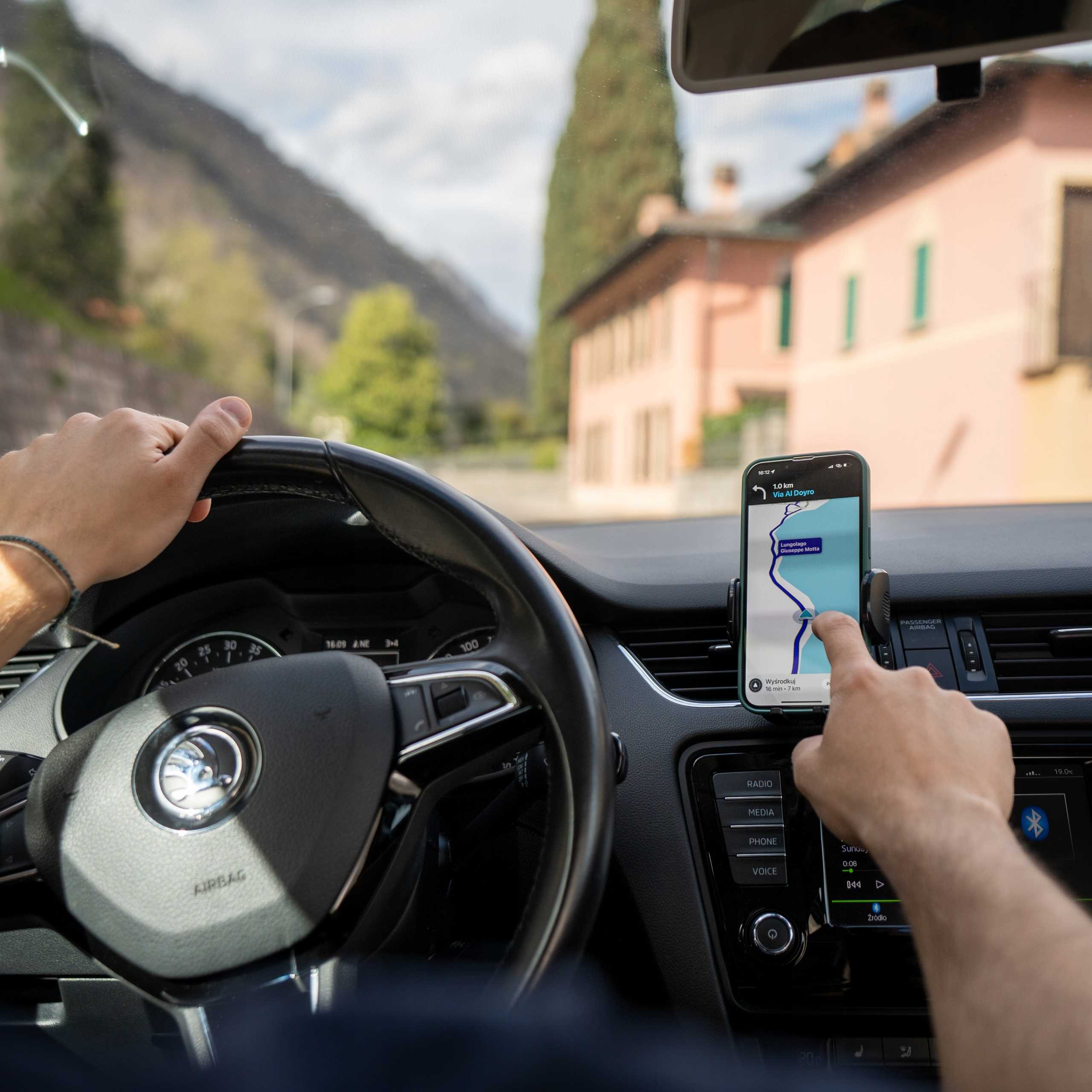 Osoba prowadzi samochód, używając telefonu do nawigacji. Budynki i góry widoczne są przez przednią szybę.