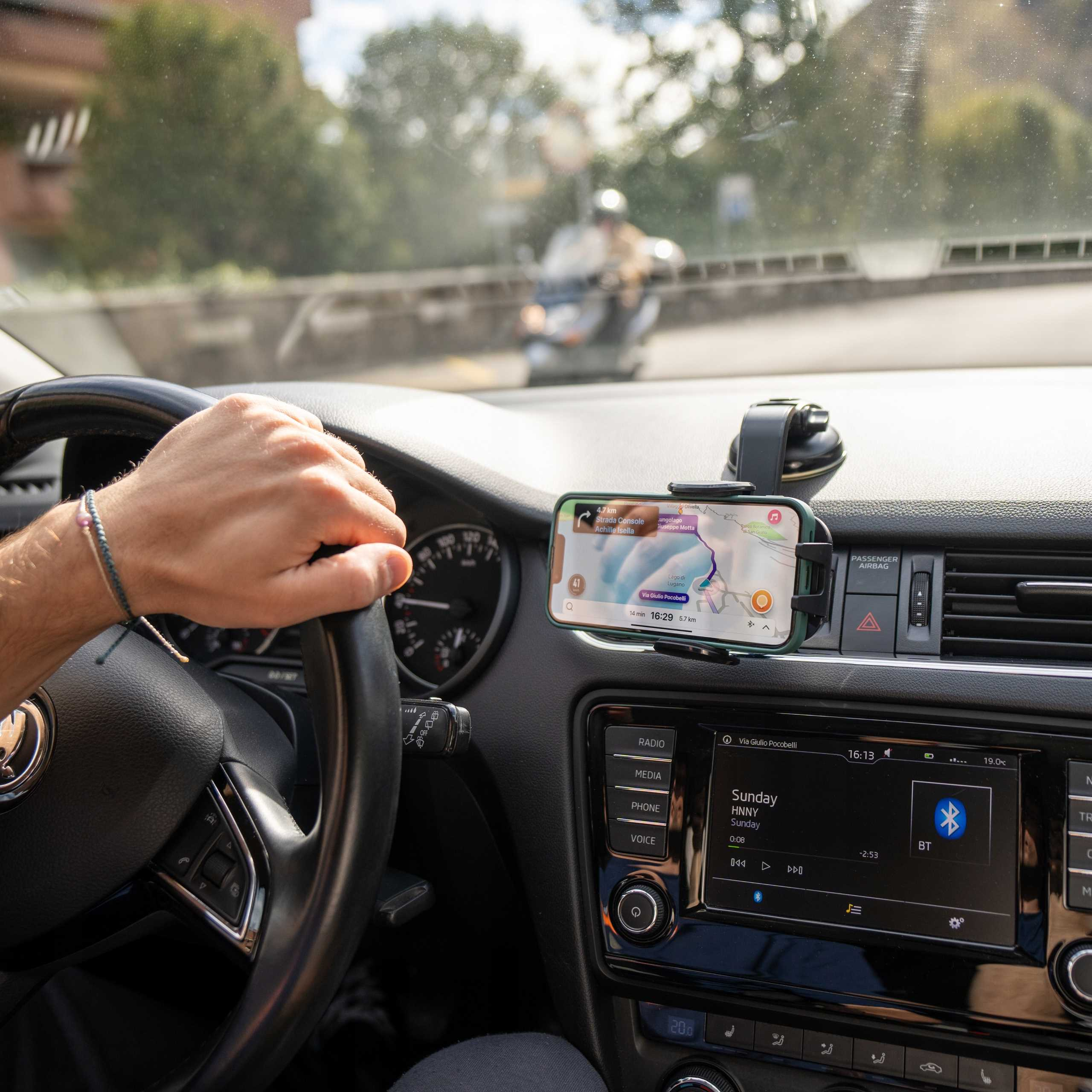 Osoba prowadzi samochód, trzymając kierownicę. Telefon wyświetla mapę. W tle widać motocykl.