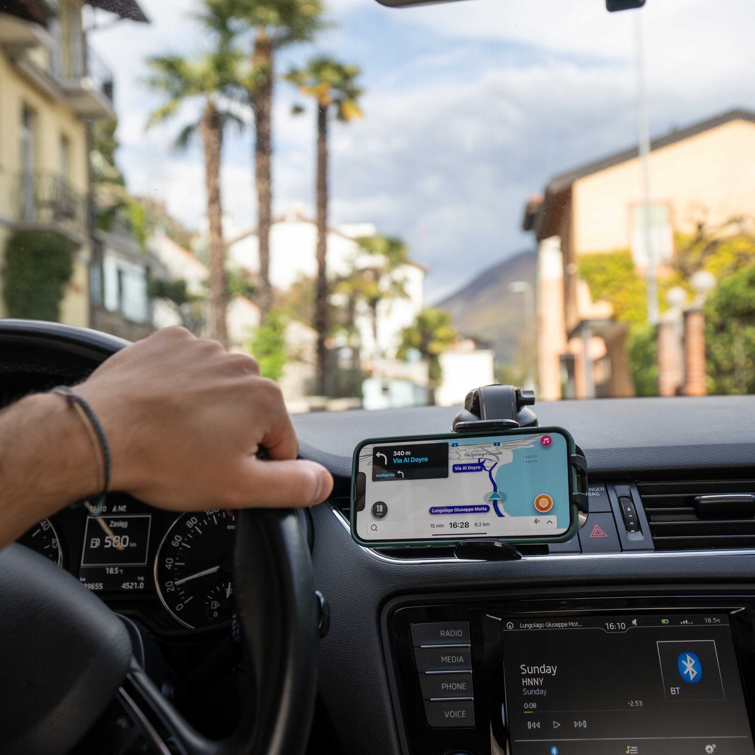 Osoba prowadzi samochód, używając telefonu z aplikacją nawigacyjną. Palmy i budynki w tle.