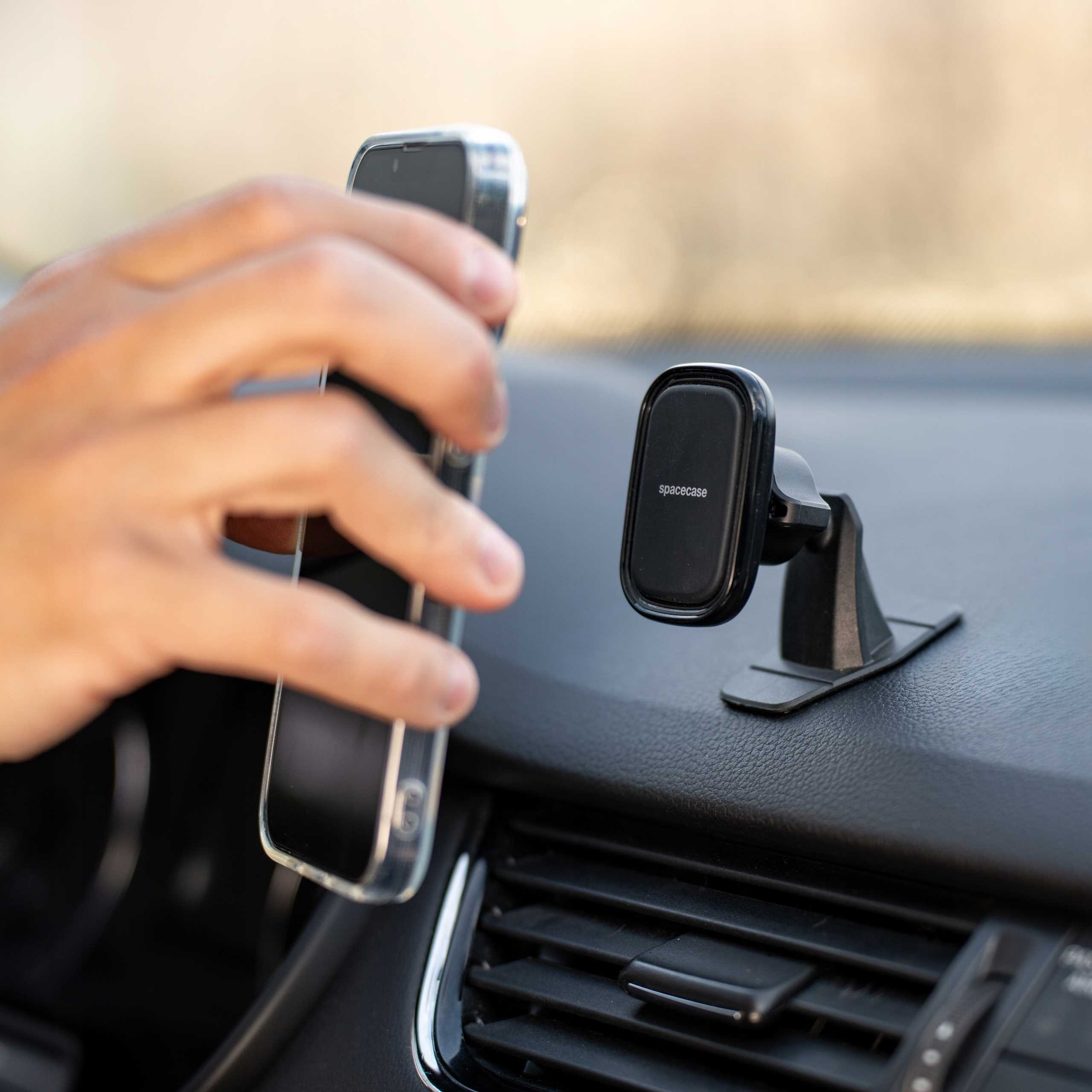 Ręka umieszcza smartfon w pobliżu czarnego uchwytu samochodowego. Uchwyt ma napis 'spacecase'. Tło to deska rozdzielcza samochodu.