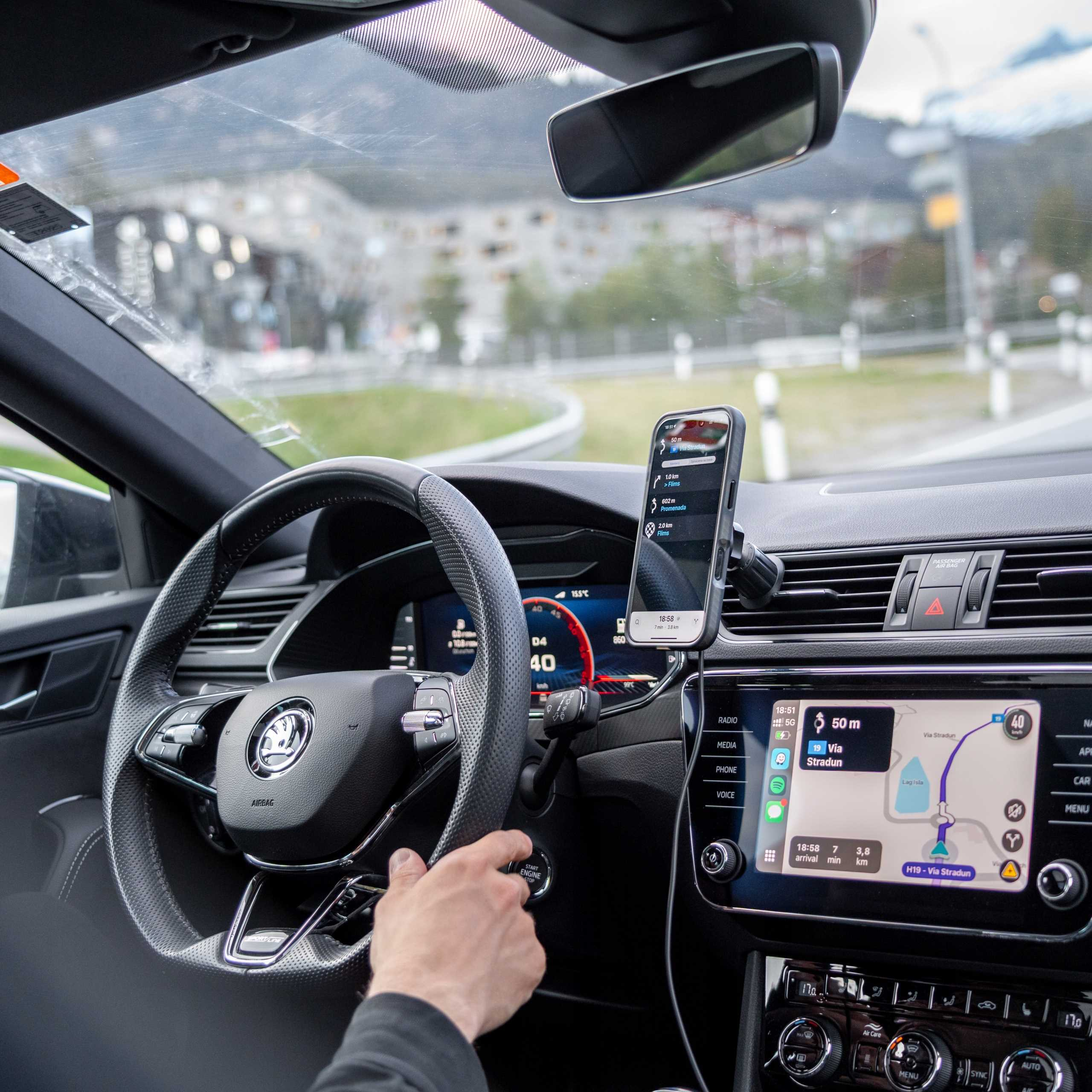 Wnętrze samochodu pokazuje rękę na kierownicy. Telefon wyświetla mapę nawigacji. Droga widoczna przez przednią szybę.