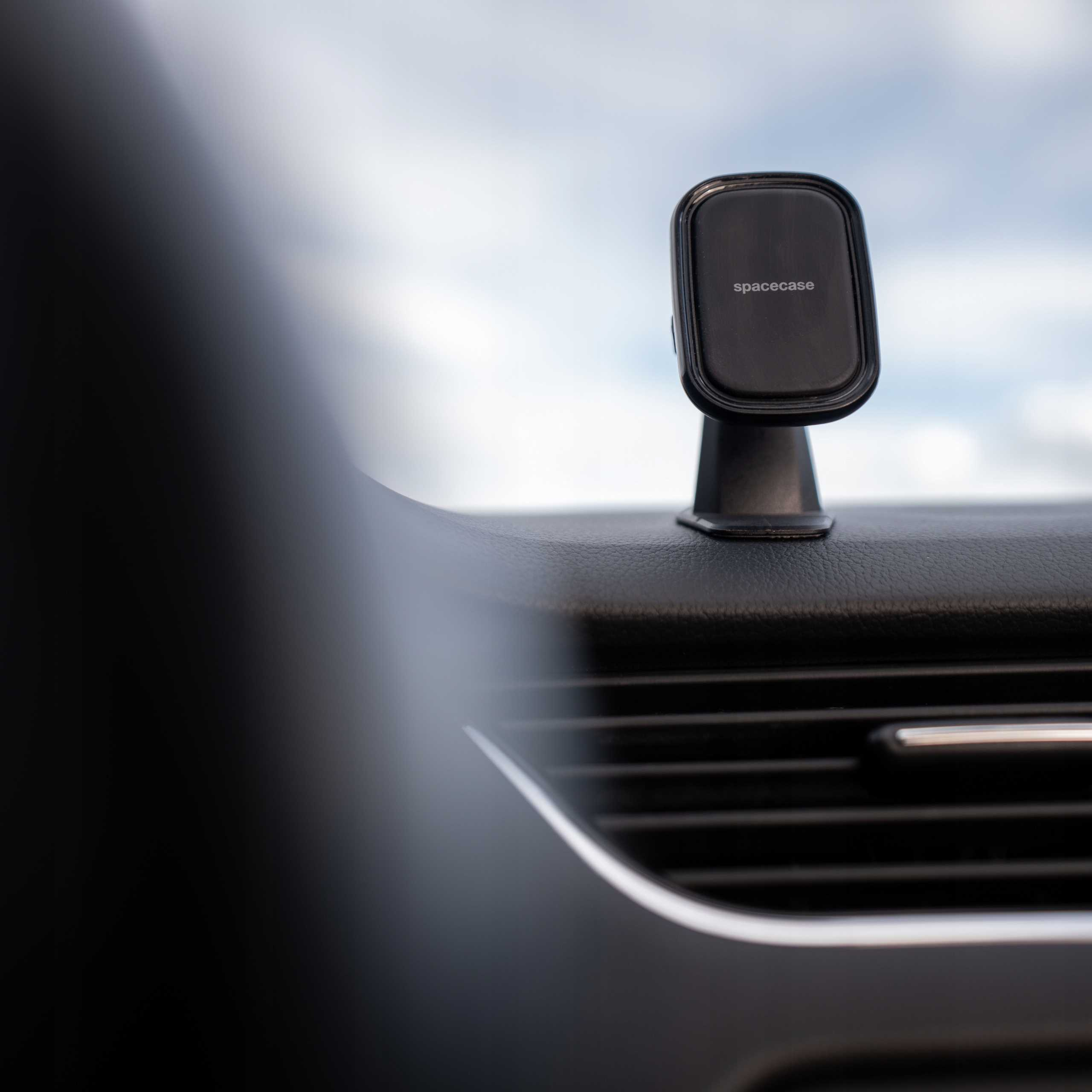 Czarny uchwyt na telefon z napisem 'spacecase' zamontowany na desce rozdzielczej samochodu. Tło to zachmurzone niebo.