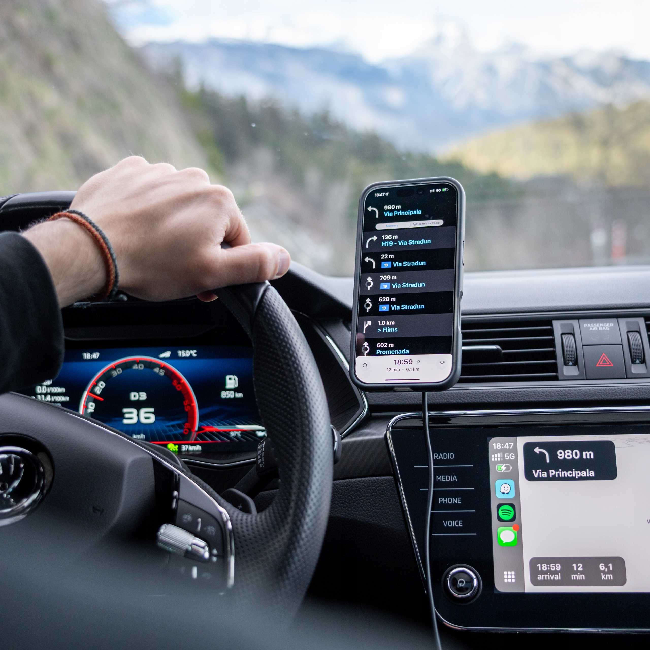 Ręka na kierownicy, trzymająca telefon z nawigacją. Deska rozdzielcza pokazuje prędkość i inne dane.