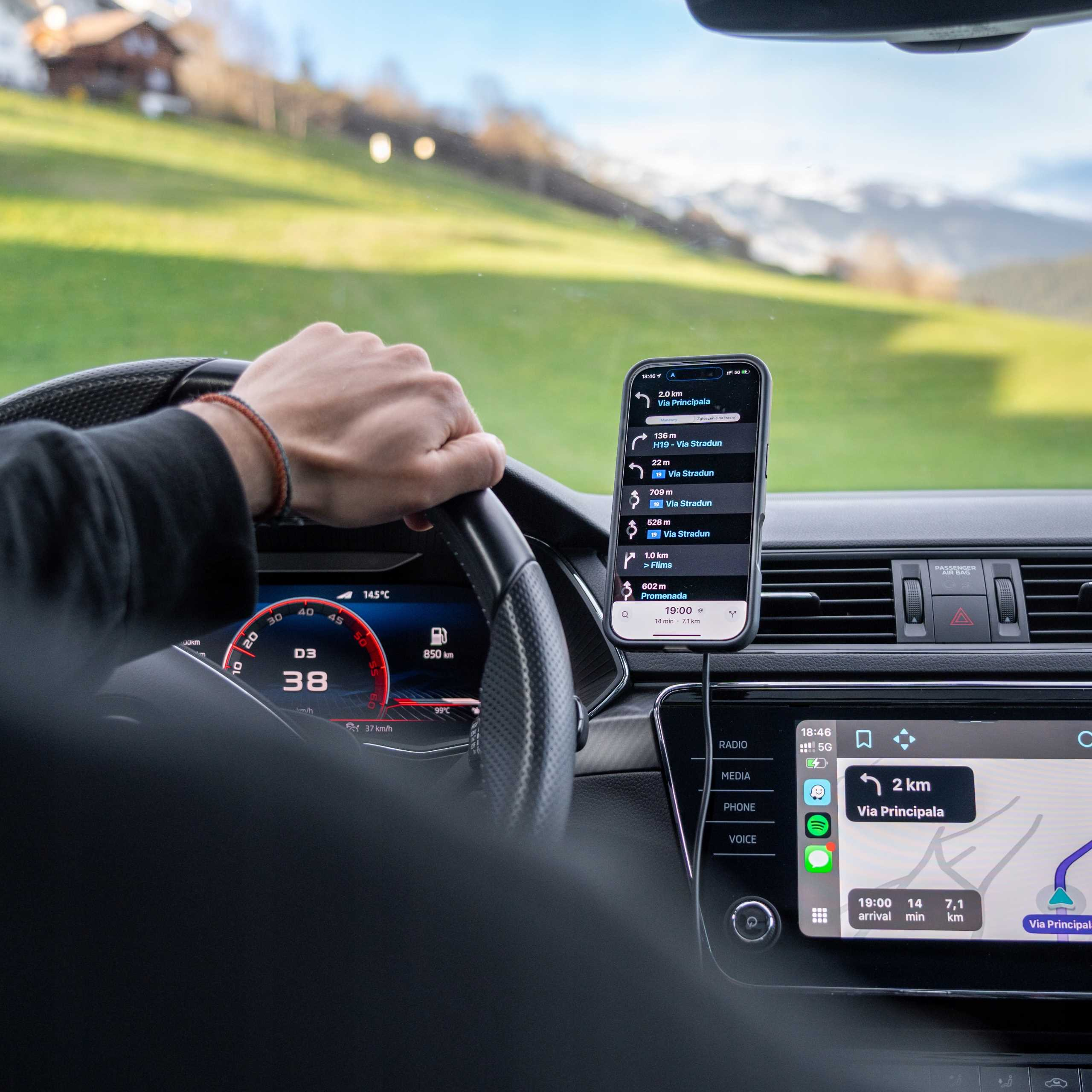Deska rozdzielcza samochodu z telefonem pokazującym nawigację. Ręka na kierownicy. Zielone wzgórza widoczne przez przednią szybę.